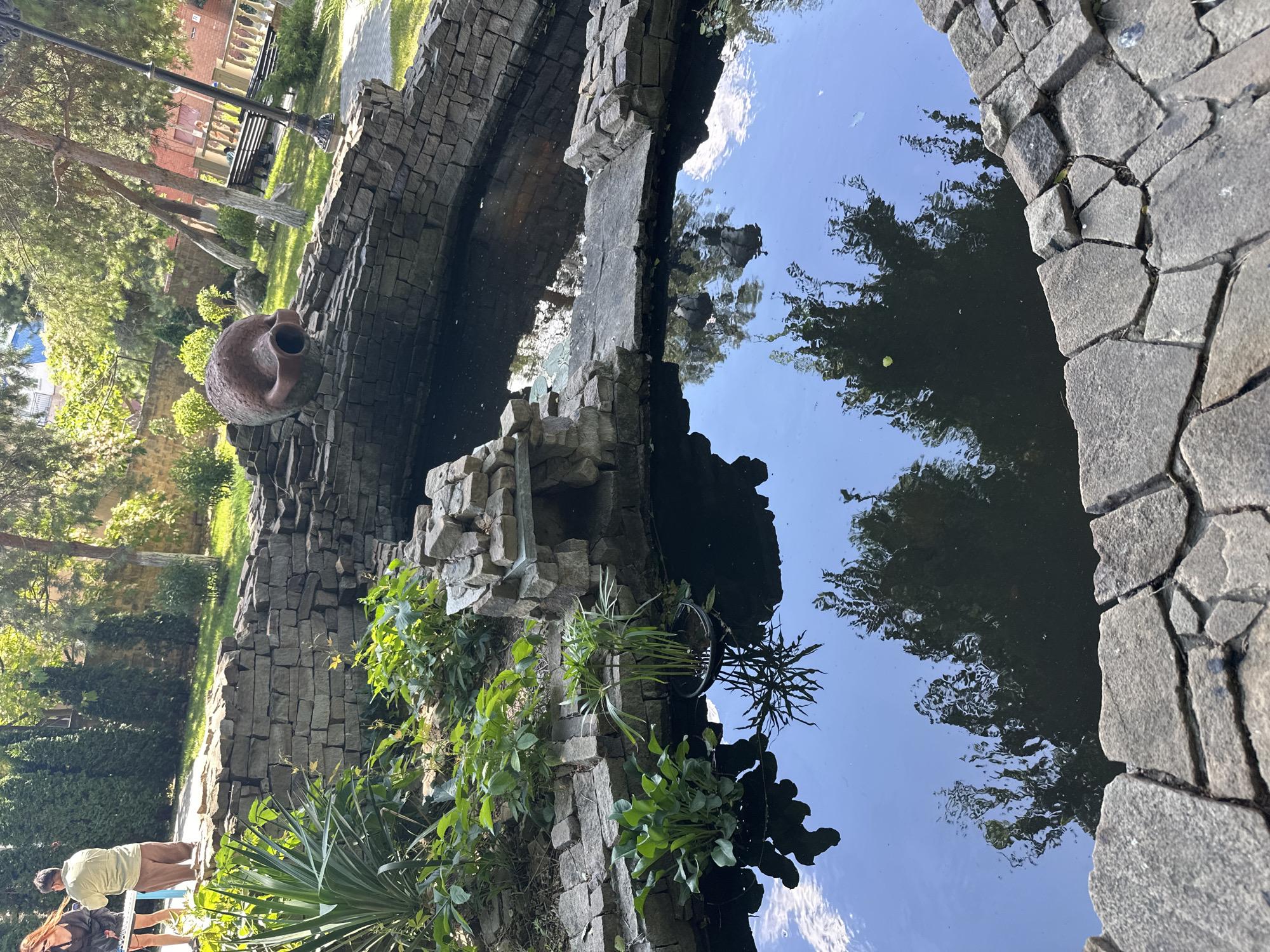 The image depicts an outdoor scene in the city of Taman, Russia. In the foreground, a stone-lined pond reflects the sky and surrounding trees. Aquatic plants grow along the edges of the pond, and a small rock structure with an opening is situated within it. A large, weathered amphora is placed on the stone wall of the pond, seemingly as a decorative element. In the background, a paved walkway is visible, leading towards a grassy area with trees. A lamppost and a brick wall with a railing are also present. Two individuals, one wearing a light-colored shirt and brown pants and the other a dark top and a skirt, are standing near the walkway, facing away from the camera. The environment suggests a park or garden setting, possibly part of a public space or a private residence.