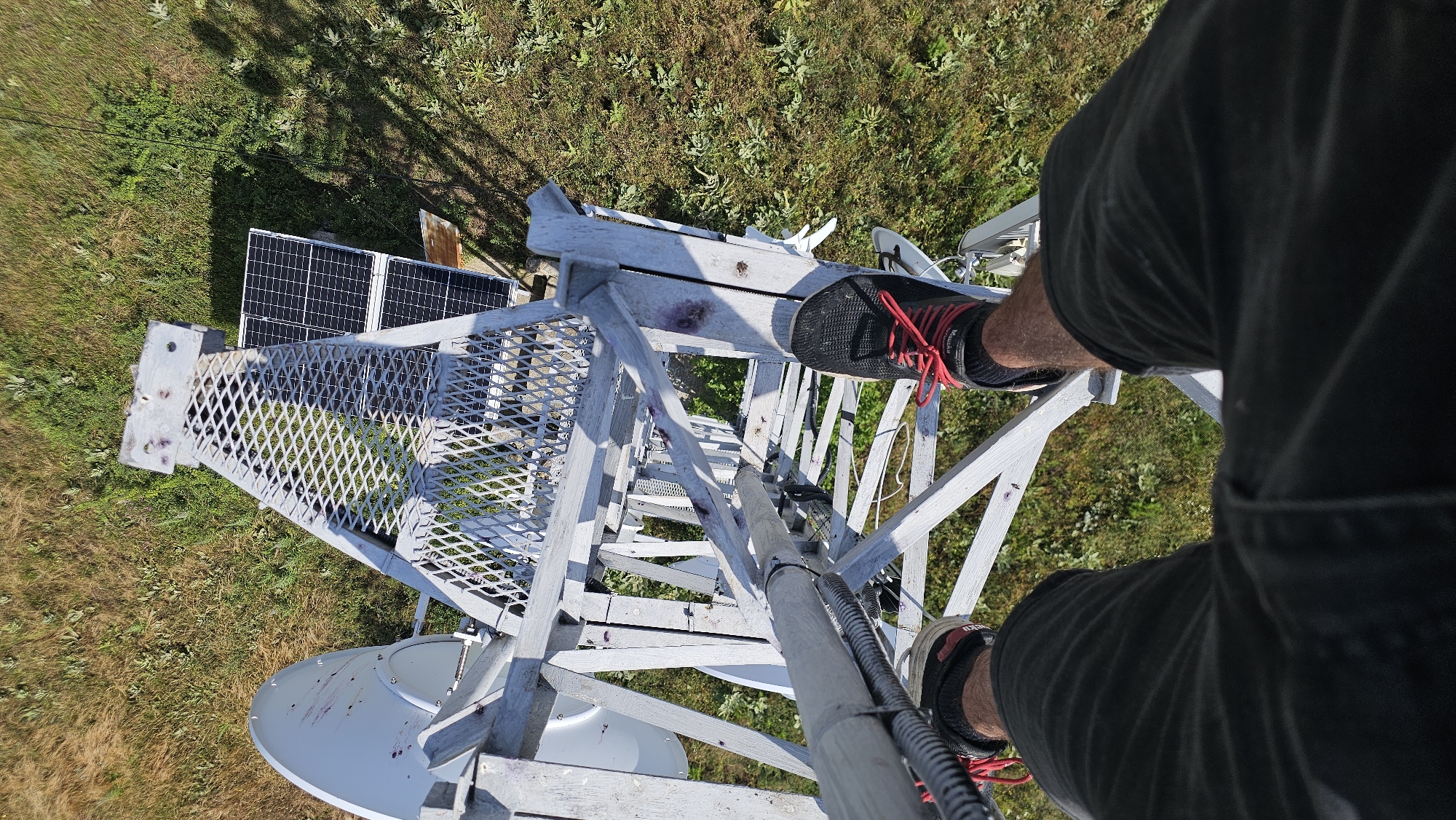 The image shows a first-person perspective of a person standing on a metal tower in Dabene, Bulgaria. The tower is made of white-painted metal with a grated walkway. Two white satellite dishes are visible attached to the side of the tower. Two solar panels are mounted to the right of the walkway, angled upwards. The ground below is covered in dry grass and green vegetation. The person is wearing black shorts and black athletic shoes with red laces. The person is standing with their feet slightly apart, looking down the tower. The scene is outdoors on a sunny day, casting shadows from the tower and vegetation.