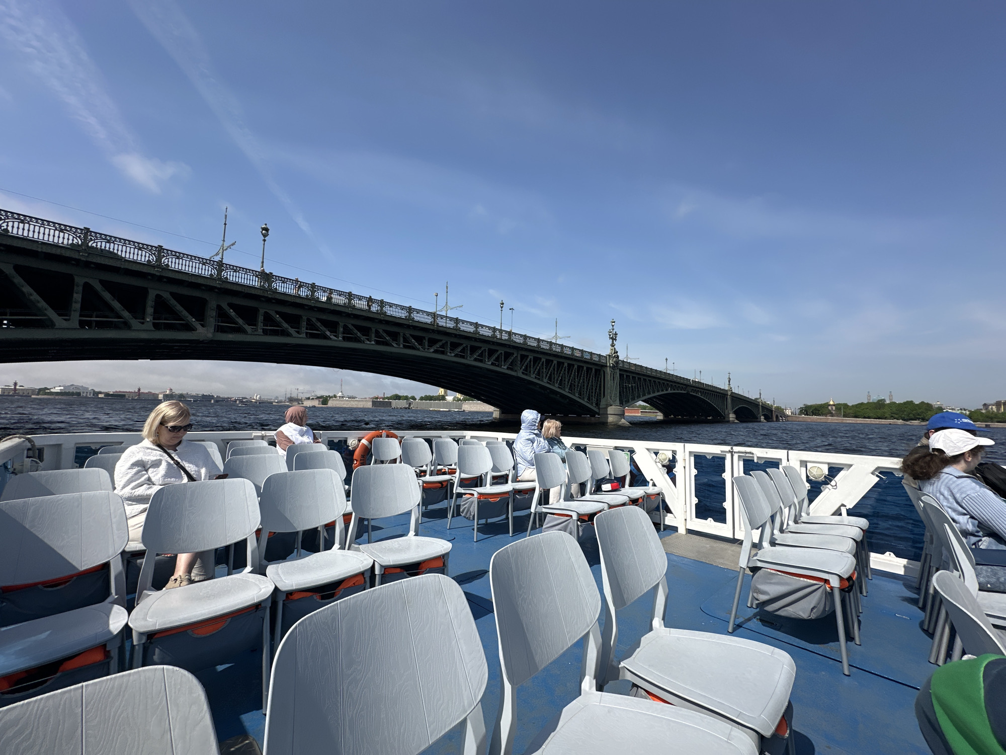 The image shows the interior of a boat traveling on a body of water, with a bridge in the background. Several rows of gray chairs are arranged on the blue deck, some occupied by passengers. An older woman in a white jacket and sunglasses is seated near the front left, and other passengers are scattered throughout the boat. A large, dark-colored bridge spans the waterway behind the boat, with buildings and greenery visible on the distant shore. The sky is blue with some wispy clouds. The location is likely Saint Petersburg, Russia, given the architectural style of the bridge and the overall urban setting.