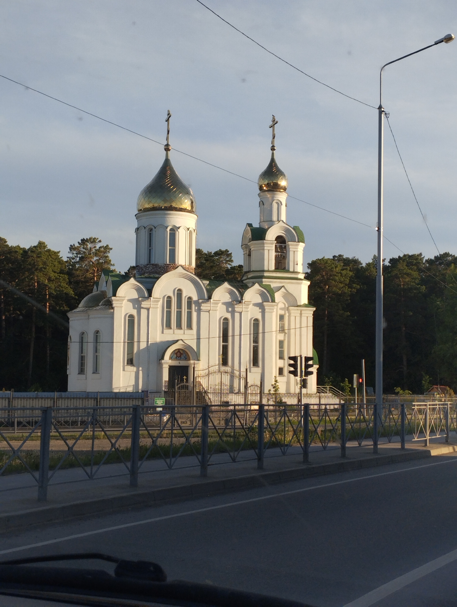 The image shows a white Orthodox church with two golden domes and crosses in Ordynskoye, Russia. The church is surrounded by a metal fence and green trees. A street is in the foreground with a lamppost and traffic lights visible. The sky is light blue with some clouds. The image was likely taken from inside a vehicle.