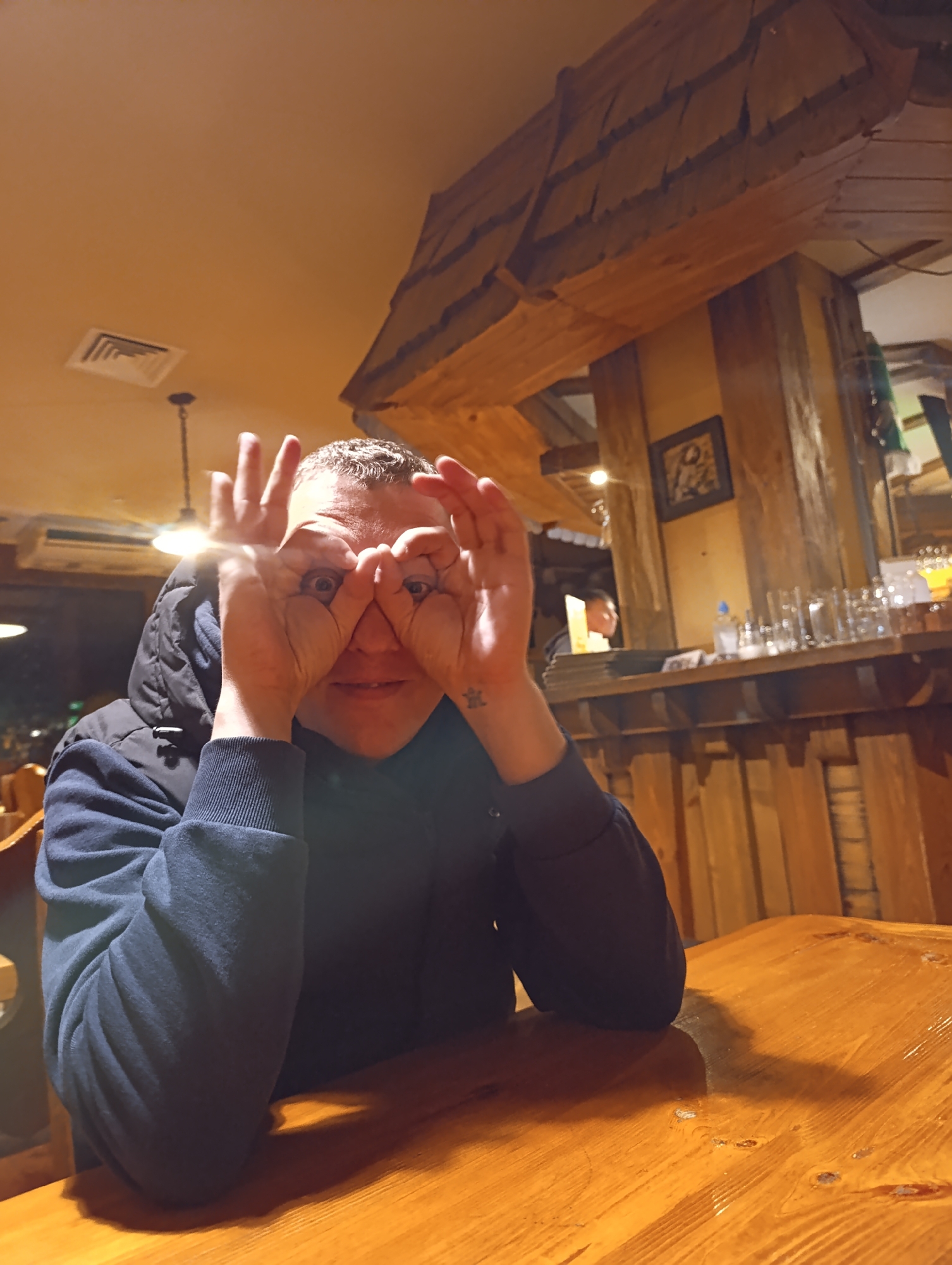 A man sits at a wooden table in what appears to be a restaurant or pub. He wears a dark blue hooded jacket. He's holding his hands up to his eyes, making a binocular-like shape with his fingers. The restaurant's interior has wooden beams and walls. In the background, a bar area can be seen with glasses and another individual behind the counter. The lighting is warm and slightly dim. There is no specific information indicating this is in Chernogolovka, Russia.