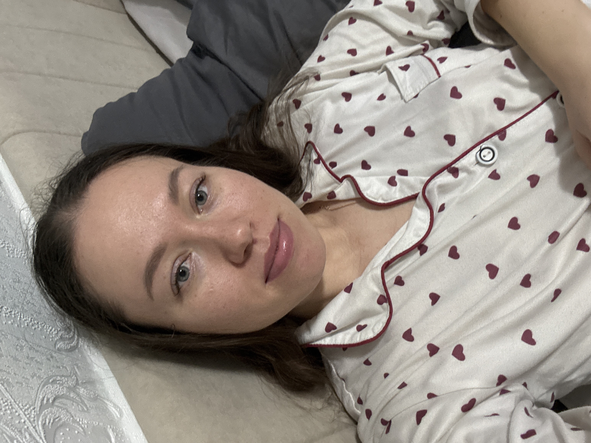 A woman with brown hair lies on a bed. She is wearing white pajamas with red hearts. She is looking at the camera. The bed is in a room, but there is no indication that it is in the city of Ферма, Russia.