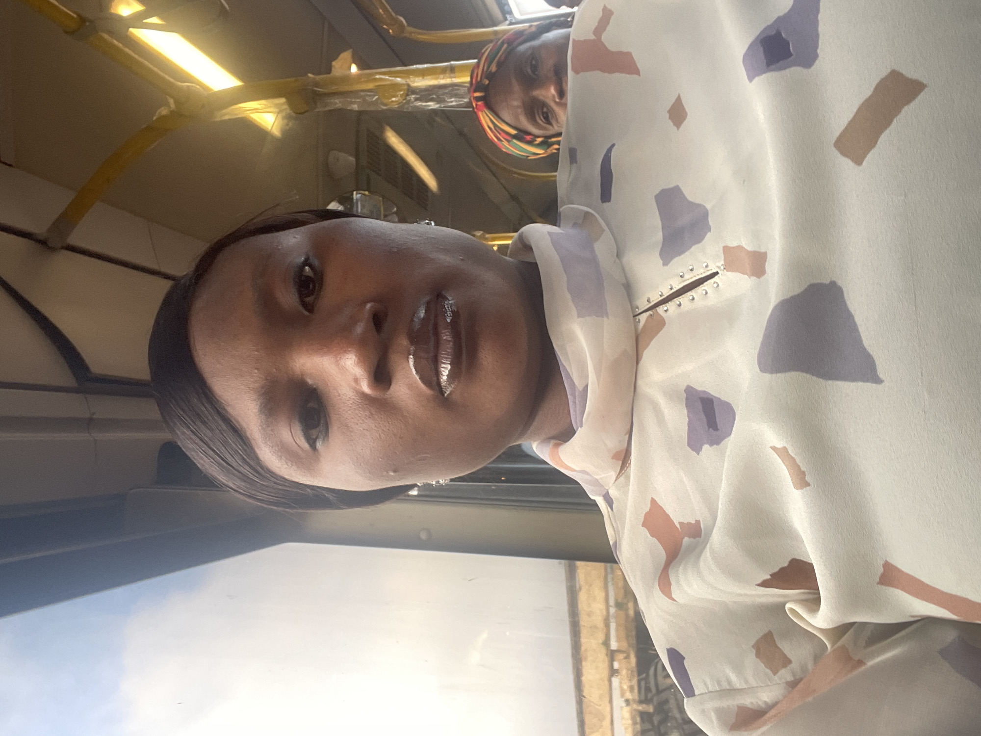 The image is a selfie taken from a bus in Jos, Nigeria. The primary subject is a woman in the foreground, looking directly at the camera. She has dark skin, dark hair styled neatly, and is wearing a light-colored blouse with a geometric pattern in shades of purple, beige, and pink. The blouse has a V-neckline with small embellishments. Her lips are glossy. In the background, another woman is partially visible, wearing a colorful headscarf. The interior of the bus is visible, with yellow poles and lighting fixtures. Outside the window, a blurred urban landscape can be seen, consistent with a city environment. The lighting suggests it is daytime.