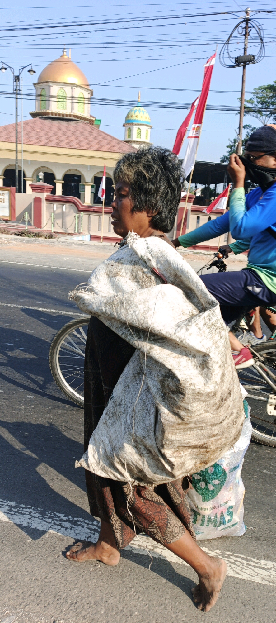 The image depicts a street scene in Kebonan, Indonesia, with a person walking barefoot across a road, carrying a large, dirty sack over their shoulder. The sack is predominantly white with brown dirt stains, and a smaller clear plastic bag with green writing and an image of what appears to be leaves or a plant is partially visible underneath it. The person is wearing a patterned skirt in earthy tones. In the background, a building with a gold dome and a smaller blue and gold dome is visible, suggesting it might be a place of worship, possibly a mosque. Indonesian flags are displayed on poles in front of the building, and a street light is also present. To the right, another person is riding a bicycle, partially obscured, wearing a blue long-sleeved shirt and dark pants, with their face covered by a dark cloth. The road has white lane markings. The lighting indicates it is daytime, casting shadows on the asphalt.