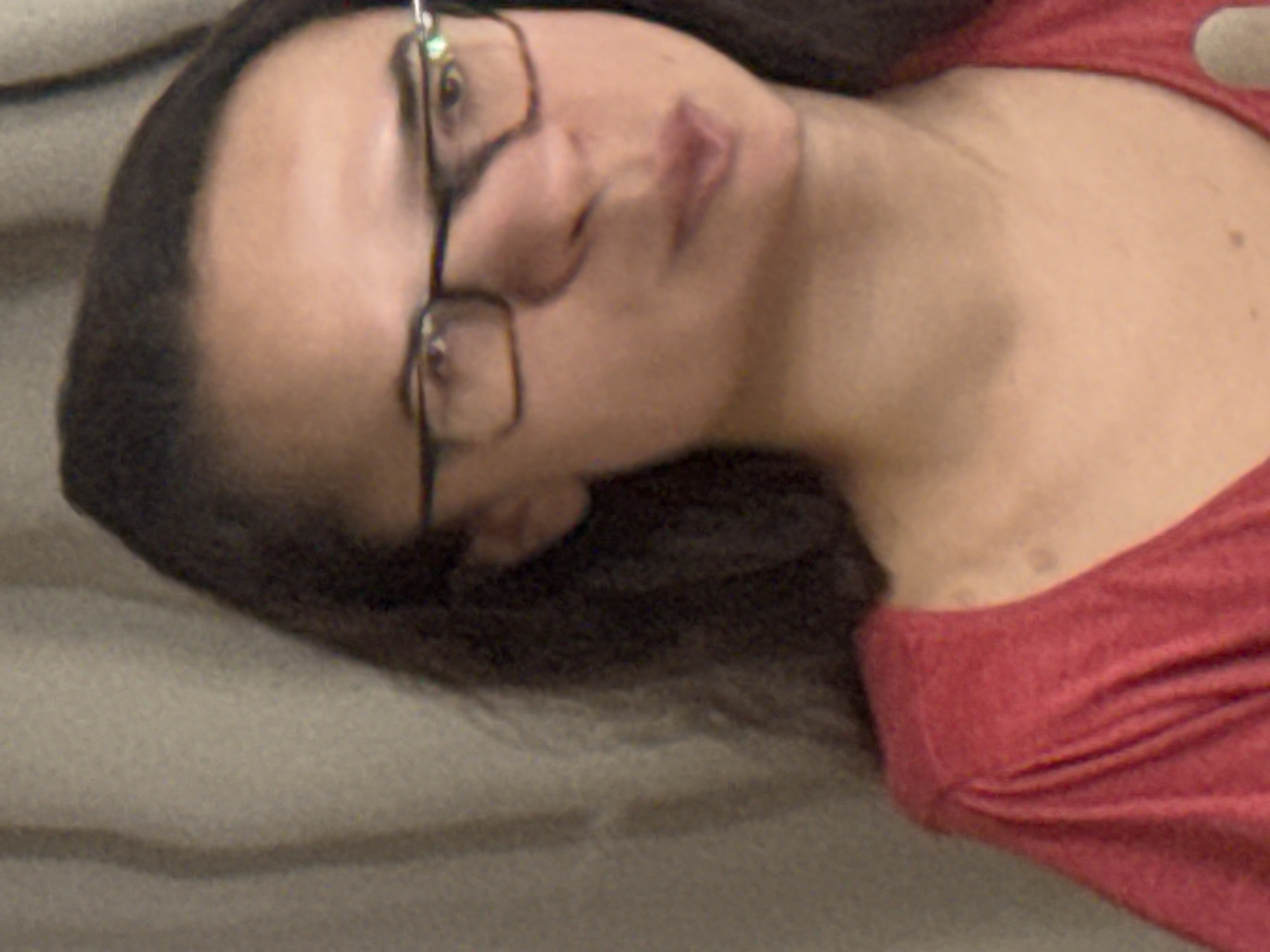 A woman with dark hair and glasses is lying down on a gray surface. She is wearing a red shirt. The location is not identifiable.