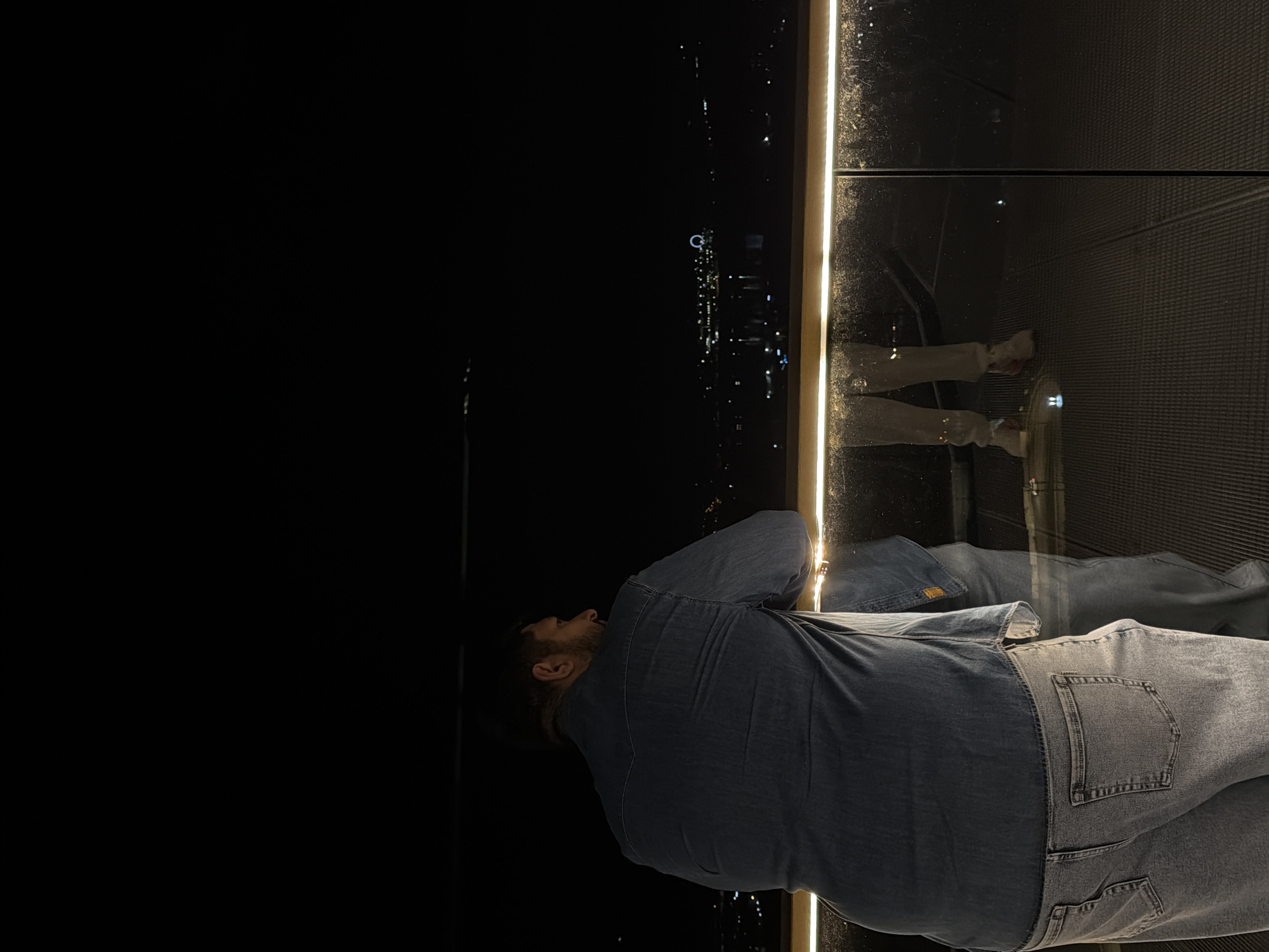 A man in a denim shirt and jeans is standing in front of a glass wall at night, looking out at the city lights. The glass wall is bordered by a narrow strip of light. The man's reflection can be seen in the glass. The city lights appear to be from Krasnoyarsk, Russia.