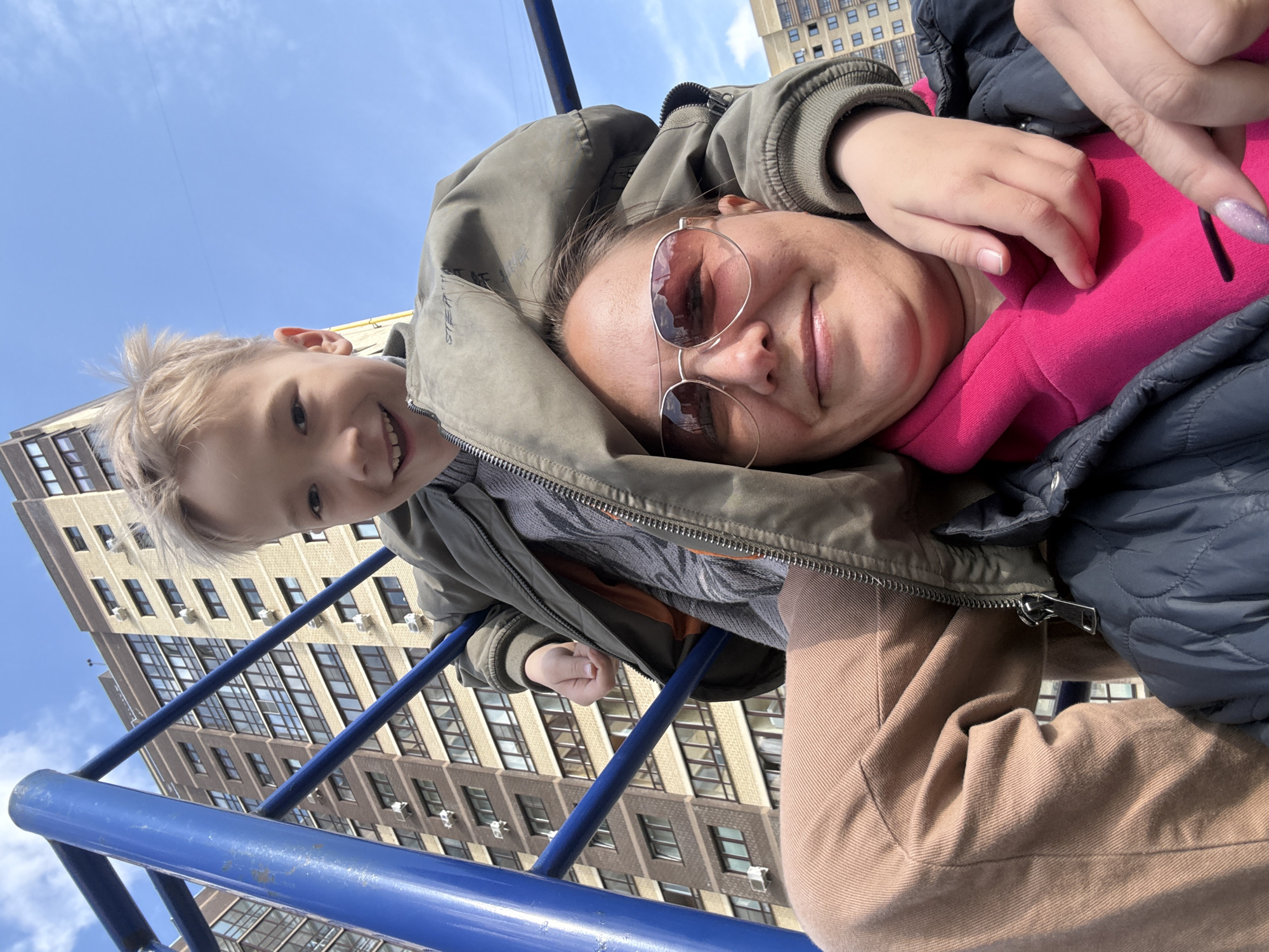 A selfie of a smiling woman wearing sunglasses and a smiling boy with blond hair and a jacket, taken at a playground in Dmitrov, Russia. The woman is wearing a pink shirt and a black jacket, and the boy is holding onto a blue metal bar. A tall building is in the background. The sky is blue with scattered clouds.