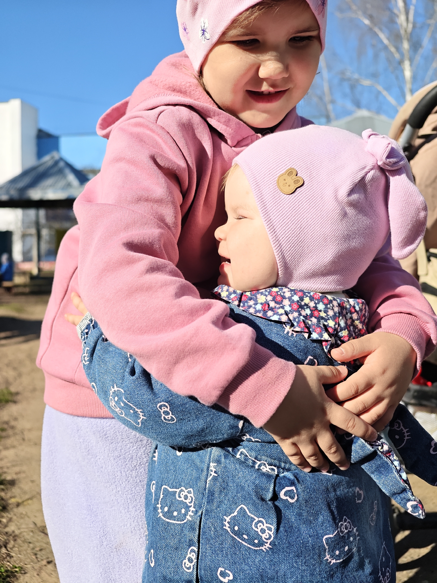 A young child in a pink hoodie and matching hat is holding a baby in a denim jacket with white cartoon characters. The baby is also wearing a pink hat. The location appears to be an outdoor area, possibly a park, in Chaikovsky, Russia. There is a building and trees in the background, and a stroller partially visible on the right side of the image.