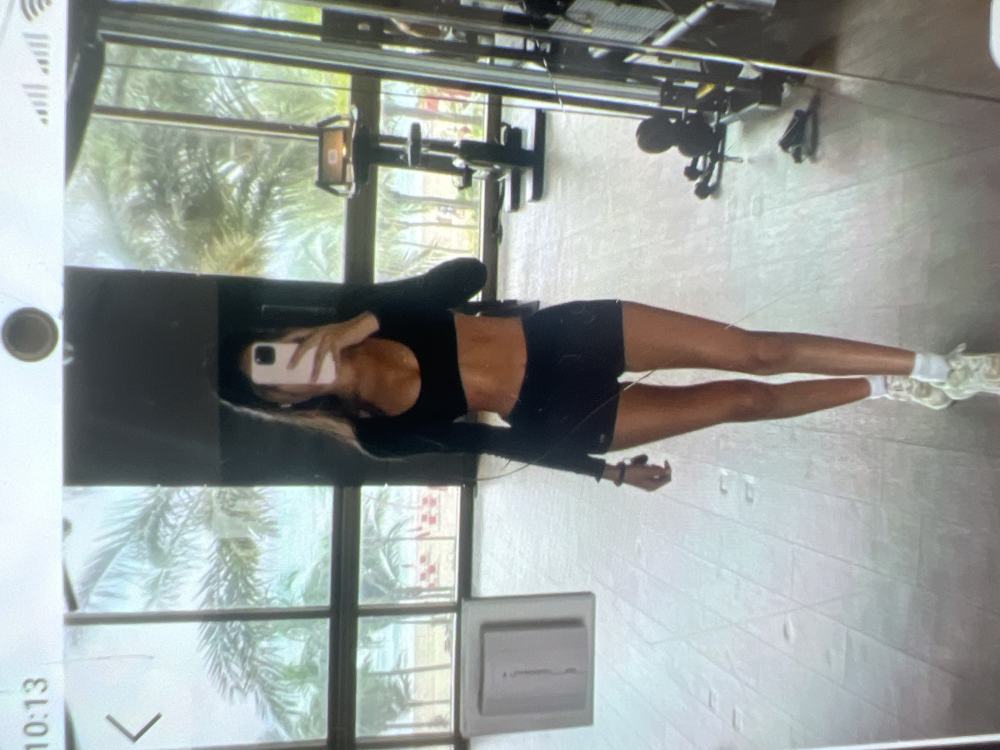 The image shows a full-body selfie taken by a woman in a gym. She is wearing a black long-sleeved crop top and black shorts. She is holding a pink smartphone in her right hand, angled to capture her reflection in a mirror. The gym appears to have large windows overlooking a tropical landscape with palm trees and a beach. Workout equipment, including a weight machine and exercise bikes, is visible in the background. The overall setting suggests an indoor fitness facility with an outdoor view. The location is not specified as Kara-Dıykan, Kyrgyzstan; the description of the environment is based solely on the visual cues in the image.