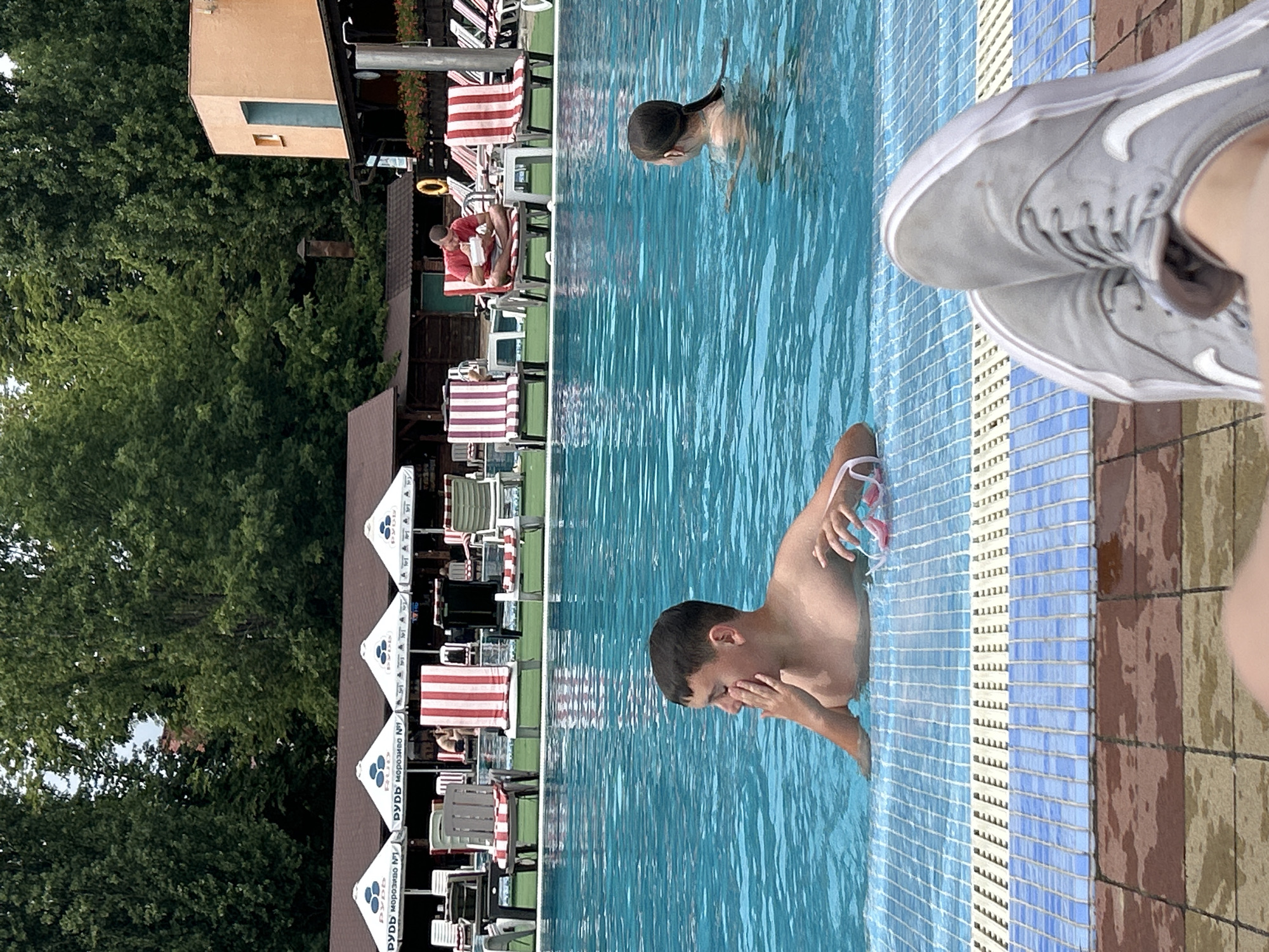 In Uzhhorod, Ukraine, a swimming pool area on a cloudy day shows several people enjoying the water and loungers. In the foreground, a person's legs wearing gray sneakers are visible, suggesting they are seated on the pool deck. The pool itself is filled with clear blue water. A young boy in the pool is touching his face. Further back, a girl with dark hair is also in the water. Around the pool, lounge chairs with red and white striped cushions are set up, some occupied. Behind the chairs, there are buildings with brown roofs and a colorful orange structure. Umbrellas with logos are providing shade. Trees with lush green foliage surround the area. The scene appears to be a public or private recreational facility.