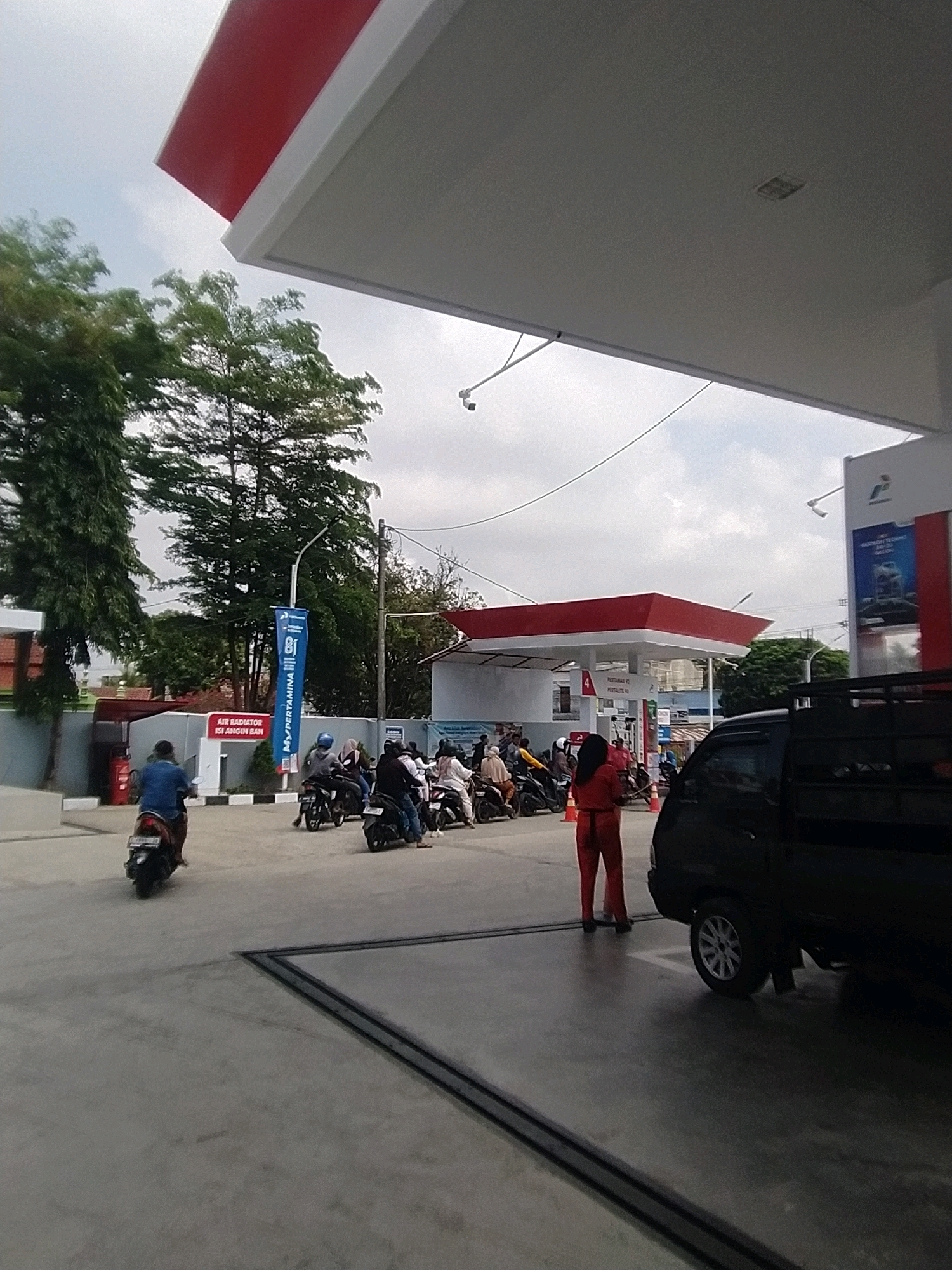 The image shows a gas station in Desa Karangbenda, Indonesia. In the foreground, a black utility vehicle is parked on the right. A person wearing a red uniform stands near it, facing away from the camera. In the middle ground, several motorcycles are lined up, with riders on them. One person on a motorcycle is turning away from the camera. A blue banner with "MYPERTAMINA" written on it is visible. In the background, a red and white gas station canopy is prominent, with trees and buildings beyond it. The sky is overcast.