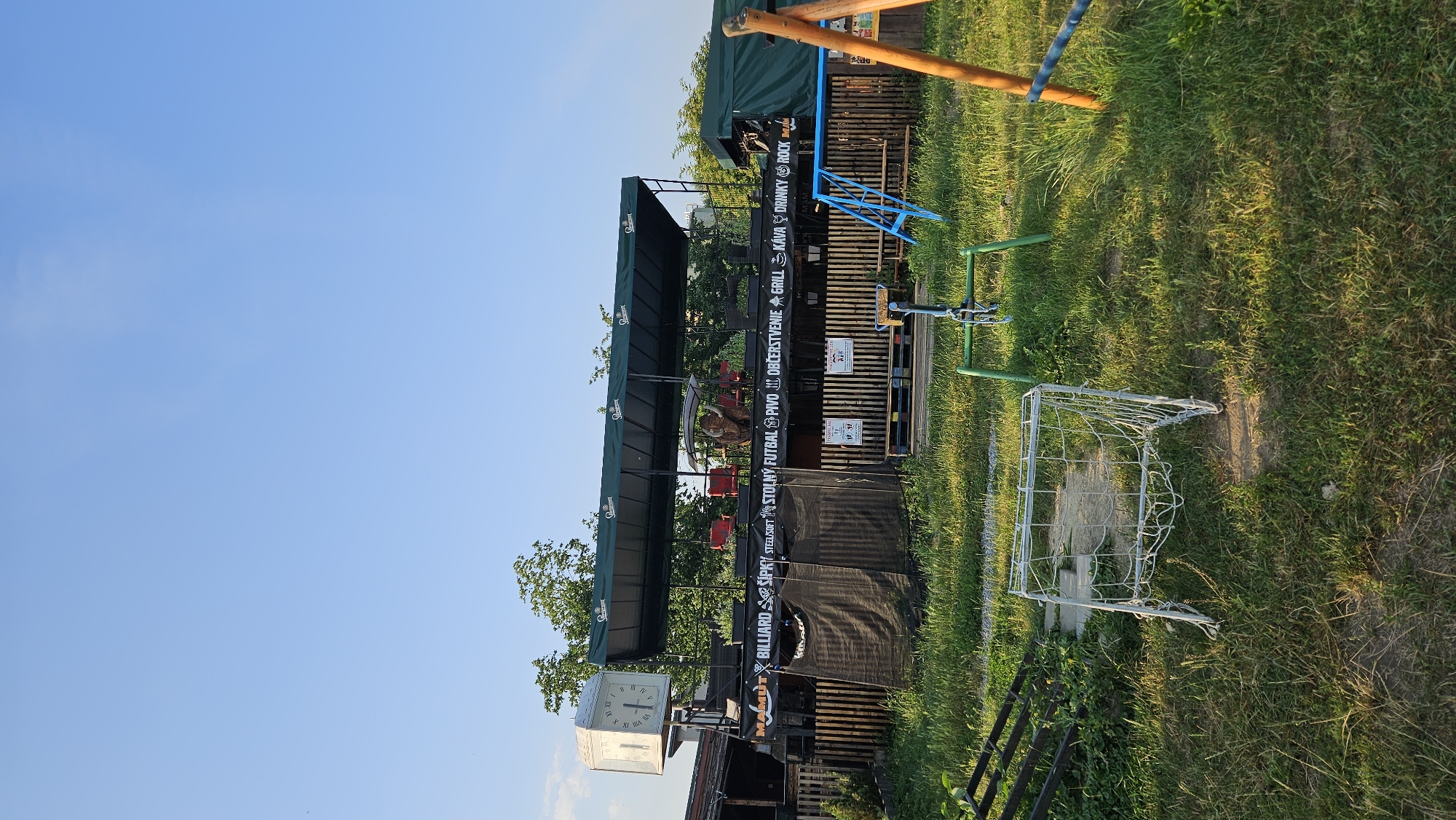 The image depicts an outdoor scene in Prešov, Slovakia, featuring a recreational area or outdoor establishment. Dominating the upper left is a large, white, square clock with Roman numerals, positioned atop a building. Below the clock and extending across the mid-ground is a covered structure with a green awning, supported by poles. Black banners with white lettering are hung below the awning, advertising various activities and offerings such as "MAMUT," "BILLIARD," "ŠÍPKY," "STEEL/SOFT," "STOLNÝ FUTBAL," "PIVO," "OBČERSTVENIE," "GRILL," "KÁVA," "DRINKY," and "ROCK." Behind this structure, a wooden fence encloses an area with tables and chairs.
In the foreground, a small, white, metal soccer goal with netting sits on a patch of grass. Further to the right, a blue metal slide structure is visible, along with parts of a wooden swing set. The ground is covered with a mixture of grass and some bare soil. The sky is clear blue, indicating fair weather. The overall impression is of a casual, outdoor entertainment venue.