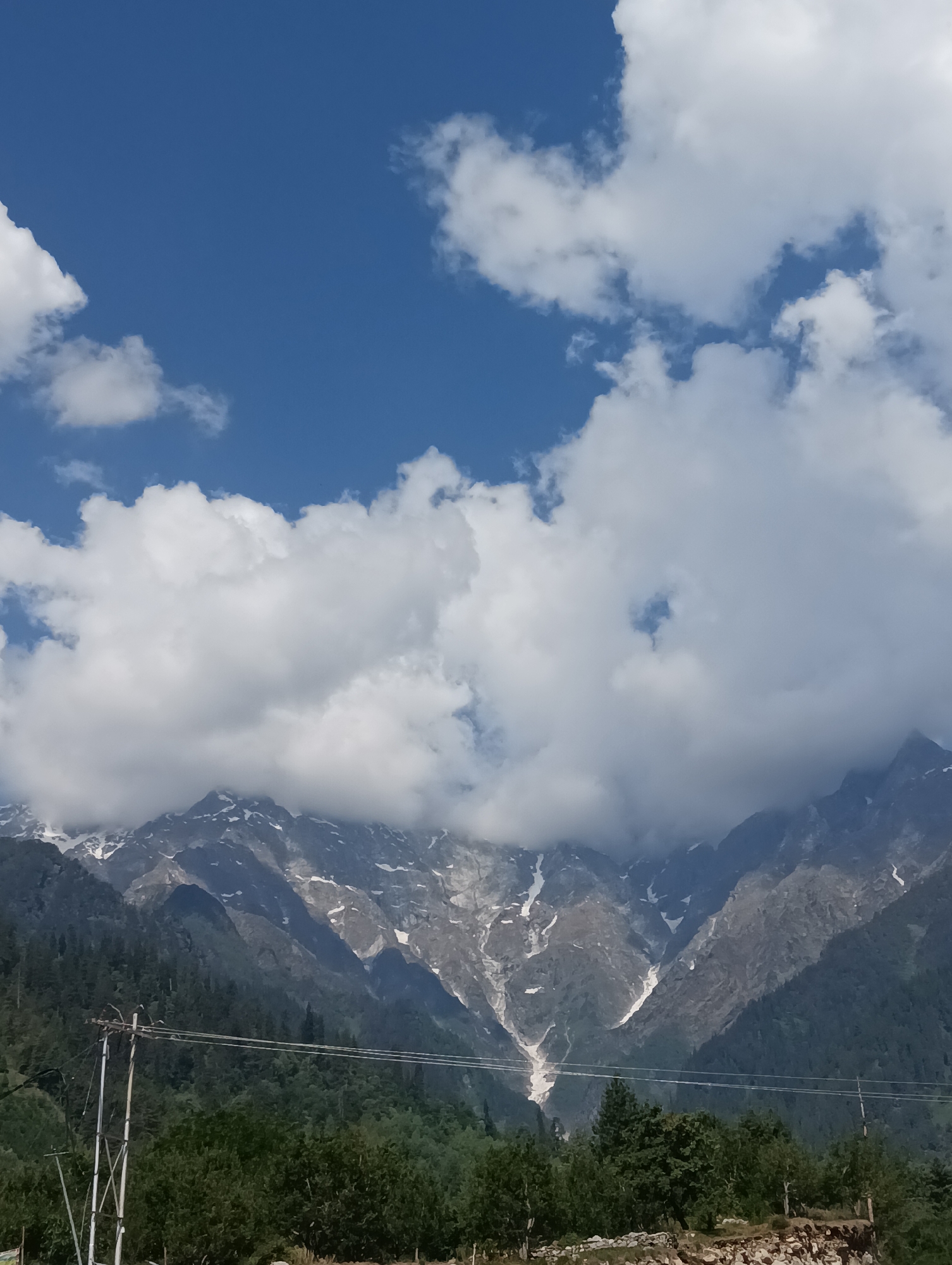 The image shows a scenic view of mountains near Palchan, India. The sky is a vibrant blue, dotted with fluffy white clouds. In the foreground, lush green trees create a dense forest that covers the lower slopes of the mountains. A power line with utility poles is visible, running horizontally across the middle of the frame. The mountains themselves are majestic, with jagged peaks partially obscured by clouds. Patches of snow can be seen on the higher elevations, indicating a cooler climate. The overall impression is one of natural beauty and tranquility.