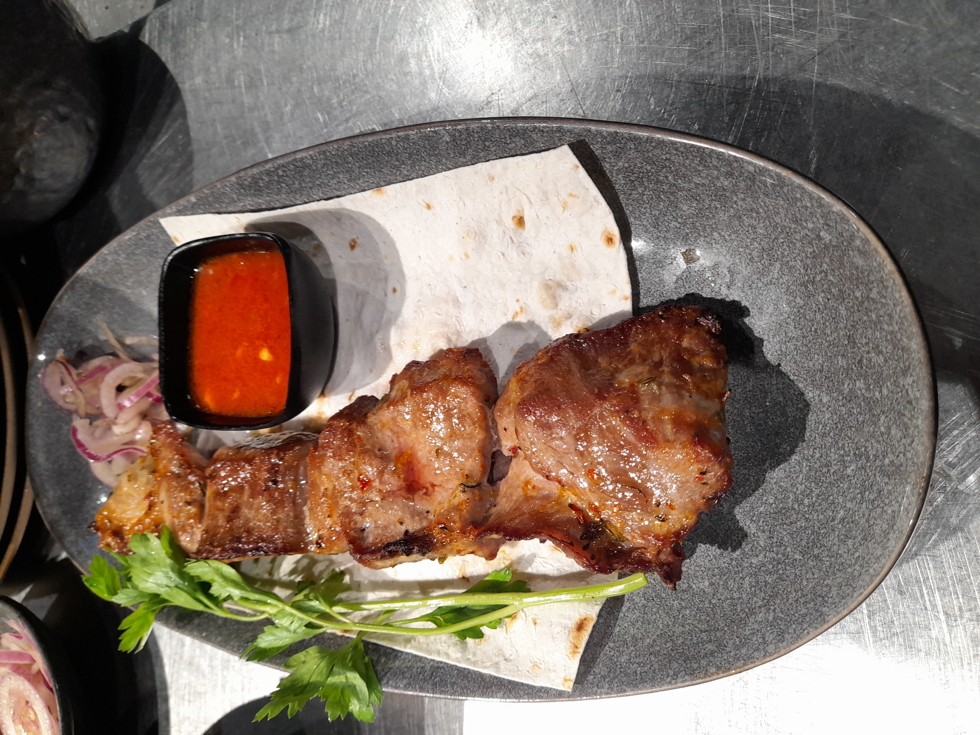 A close-up, top-down view captures a plate of grilled meat skewers served with a red sauce and pickled red onions. The skewers are arranged on a piece of flatbread, with fresh parsley garnishing the side. The plate, made of dark, textured ceramic, rests on a metallic surface. The background is blurred, hinting at a restaurant setting, possibly in Vladivostok, Russia, given the context. The lighting suggests an indoor environment, with shadows cast by the food and plate. The focus is sharp on the food, highlighting the grill marks on the meat and the vibrant color of the sauce. No people are visible in the frame.
