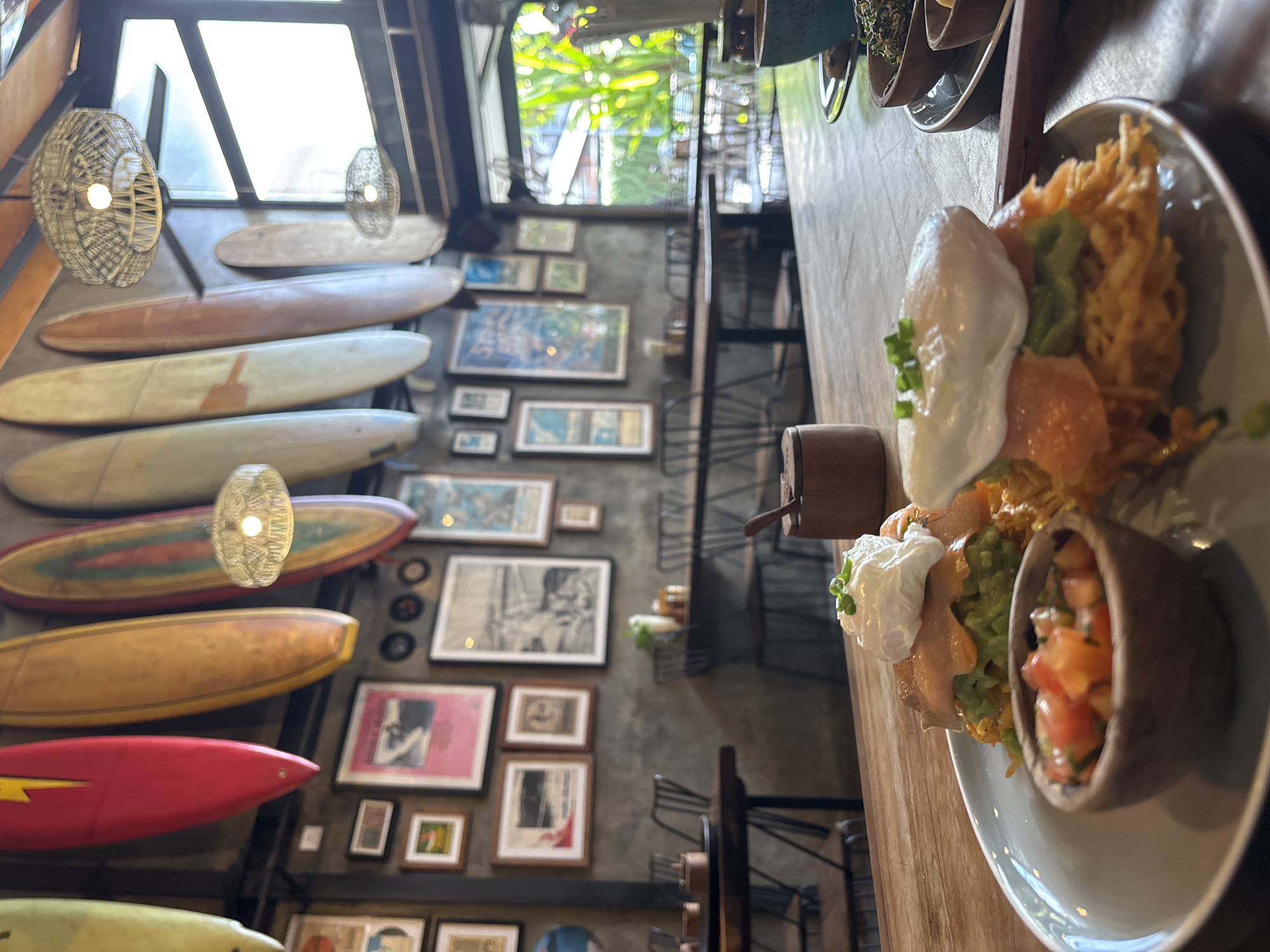 The image captures a meal at a restaurant, likely in North Kuta, Indonesia, given the decor. The focus is on a plate with a dish composed of fried strands, salmon, a dollop of white cream, guacamole, and a small bowl of salsa. Beside the plate is a small wooden container. The background is a busy interior with surfboards mounted on the wall, along with framed photos and artwork. Natural light streams in through windows. Several tables are visible, along with chairs, indicating a cafe or restaurant setting.