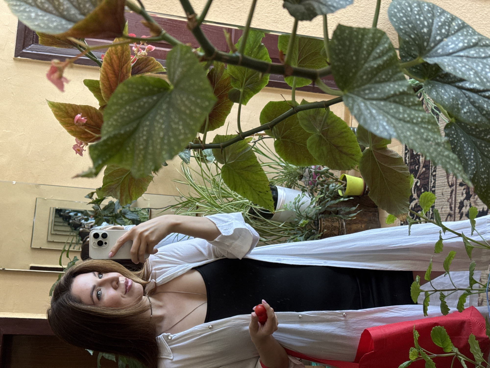 A woman with brown hair is taking a mirror selfie in what appears to be a home setting. She is wearing a black tank top under an open white button-down shirt and holding a small red object, possibly a strawberry or a charm, in her left hand. To her left, a bright red tote bag is visible. The background features several potted plants, including a large begonia with prominent, spotted leaves, and a mirror reflecting more plants and part of the room. The wall behind the woman is painted a light yellow. The overall impression is a candid moment in a well-lit, domestic environment with an abundance of greenery. This scene is consistent with an interior space in Odesa, Ukraine.