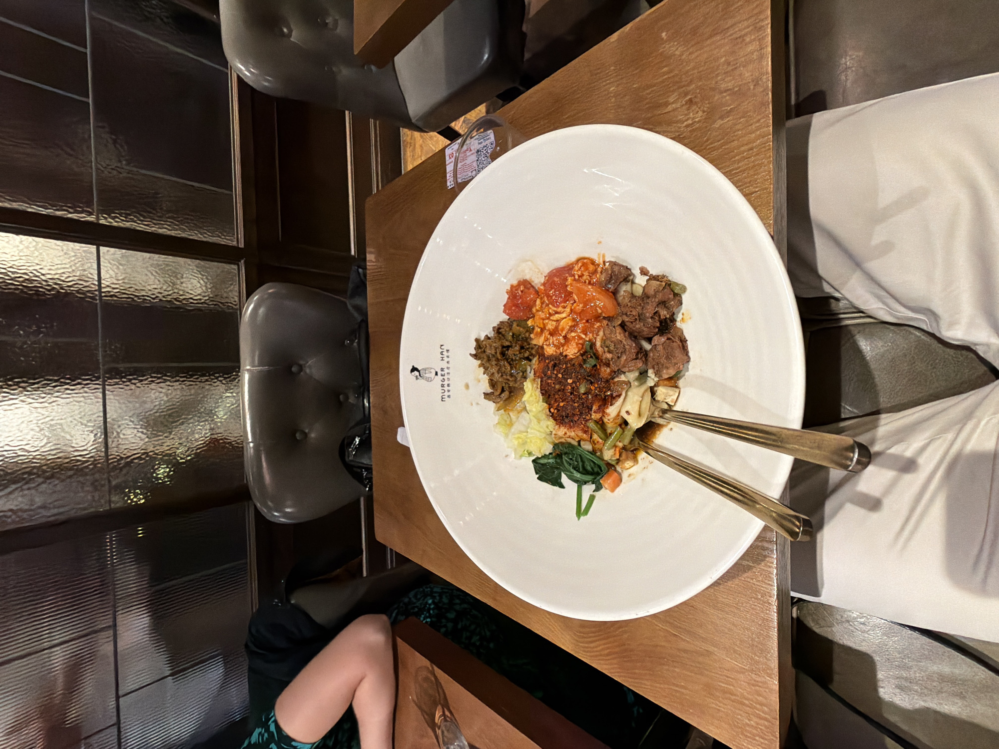 A partially seen person's legs wearing light-colored trousers are positioned in the foreground, casting a shadow. They are seated at a wooden table where a large white bowl of food is centrally placed. The bowl contains rice topped with various ingredients including chopped meat, vegetables such as spinach and carrots, and what appears to be a spicy sauce. Two golden cutlery items, likely forks or spoons, are resting across the edge of the bowl. In the background, leather chairs and a textured glass partition are visible, suggesting an indoor dining setting, possibly a restaurant in London, United Kingdom.