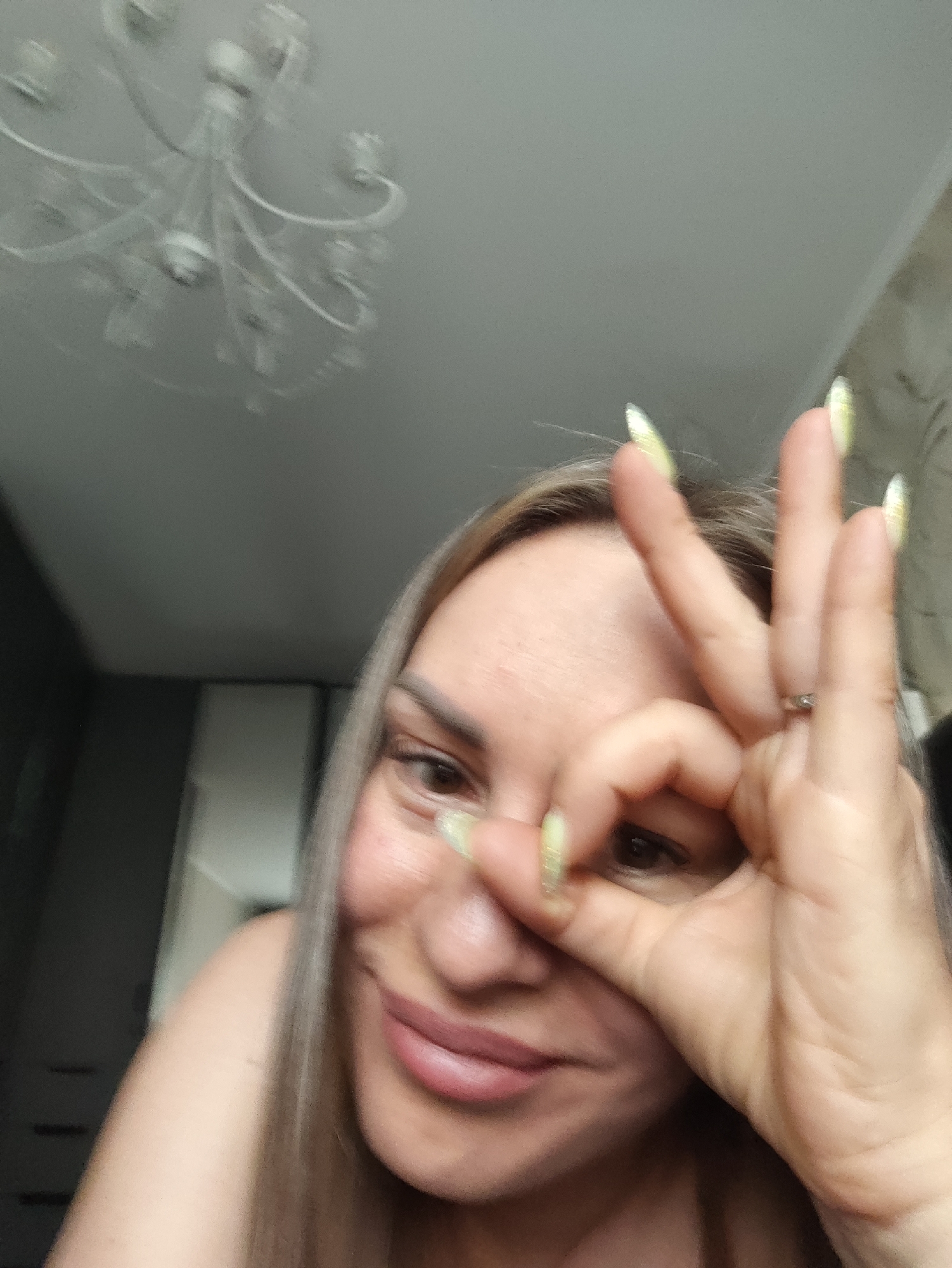 This media file shows a close-up of a person's face. The subject has light brown hair and is looking at the camera. Their right hand is raised to their face, with the index finger and thumb forming a circle around their right eye. The person is wearing a ring on their left ring finger and has light-colored fingernails. Their lips are slightly parted, suggesting a smile or a playful expression. In the background, a blurred chandelier is visible on the ceiling, and to the left, a dark surface possibly indicating furniture or a wall. The overall setting appears to be indoors, likely a home. The lighting is soft, and the image has a slightly grainy quality, suggesting it was taken in low light conditions or with a lower resolution camera. The image is likely a selfie, given the close-up perspective and the hand gesture. No text is visible in the image. The location context of Almaty, Kazakhstan, does not provide any specific visual cues within the image itself.