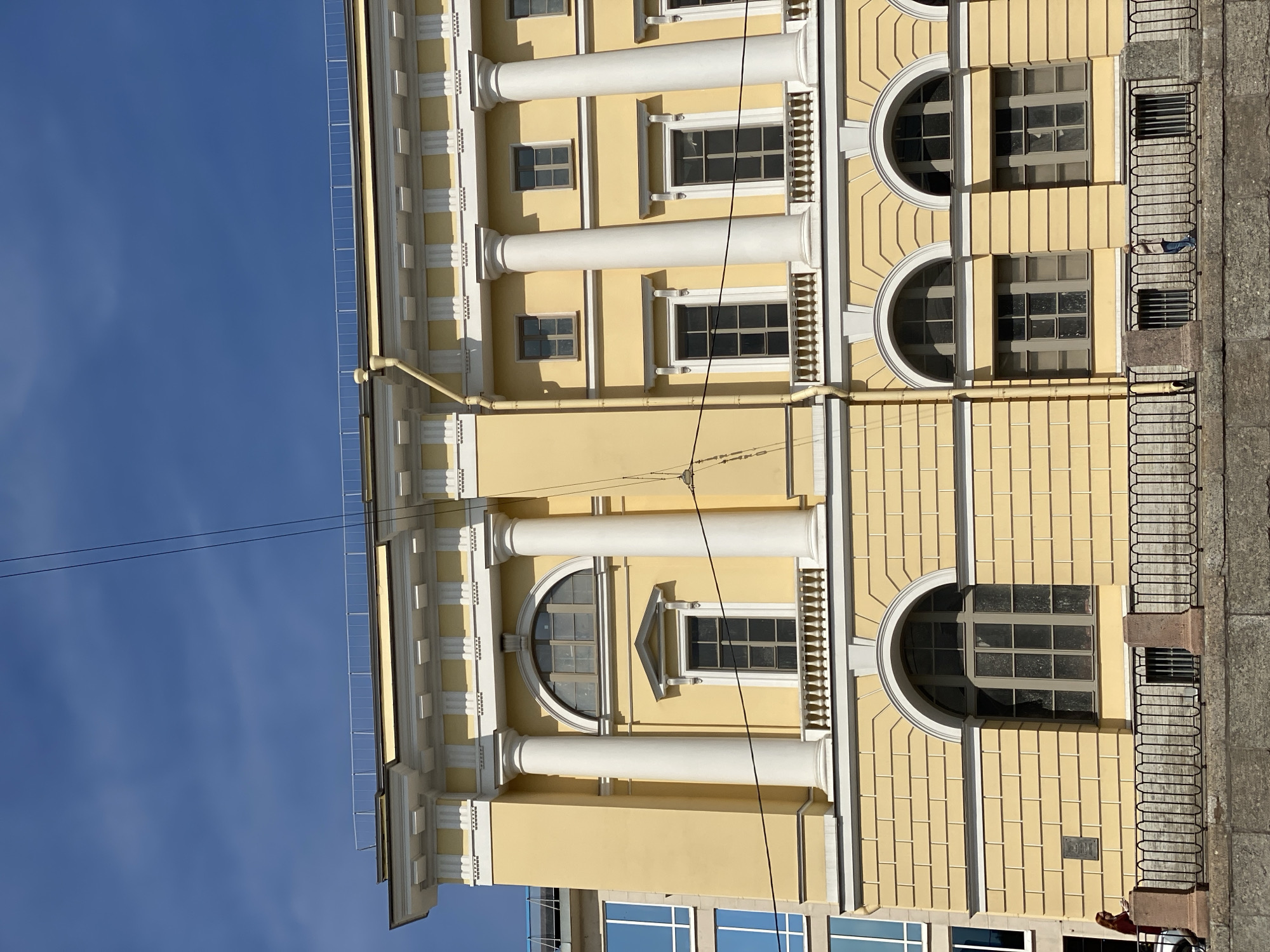 The image shows a side view of a yellow classical building with large white columns and arched windows. A railing runs along the front of the building, and a canal or river is visible in the foreground. The sky is blue with some clouds. There are two women walking along the canal. One woman is wearing a dark red jacket and the other is wearing a beige jacket. The building is located on the Moyka River Embankment in Saint Petersburg, Russia, and is part of the Marble Palace complex. The style of the building suggests it is historic.