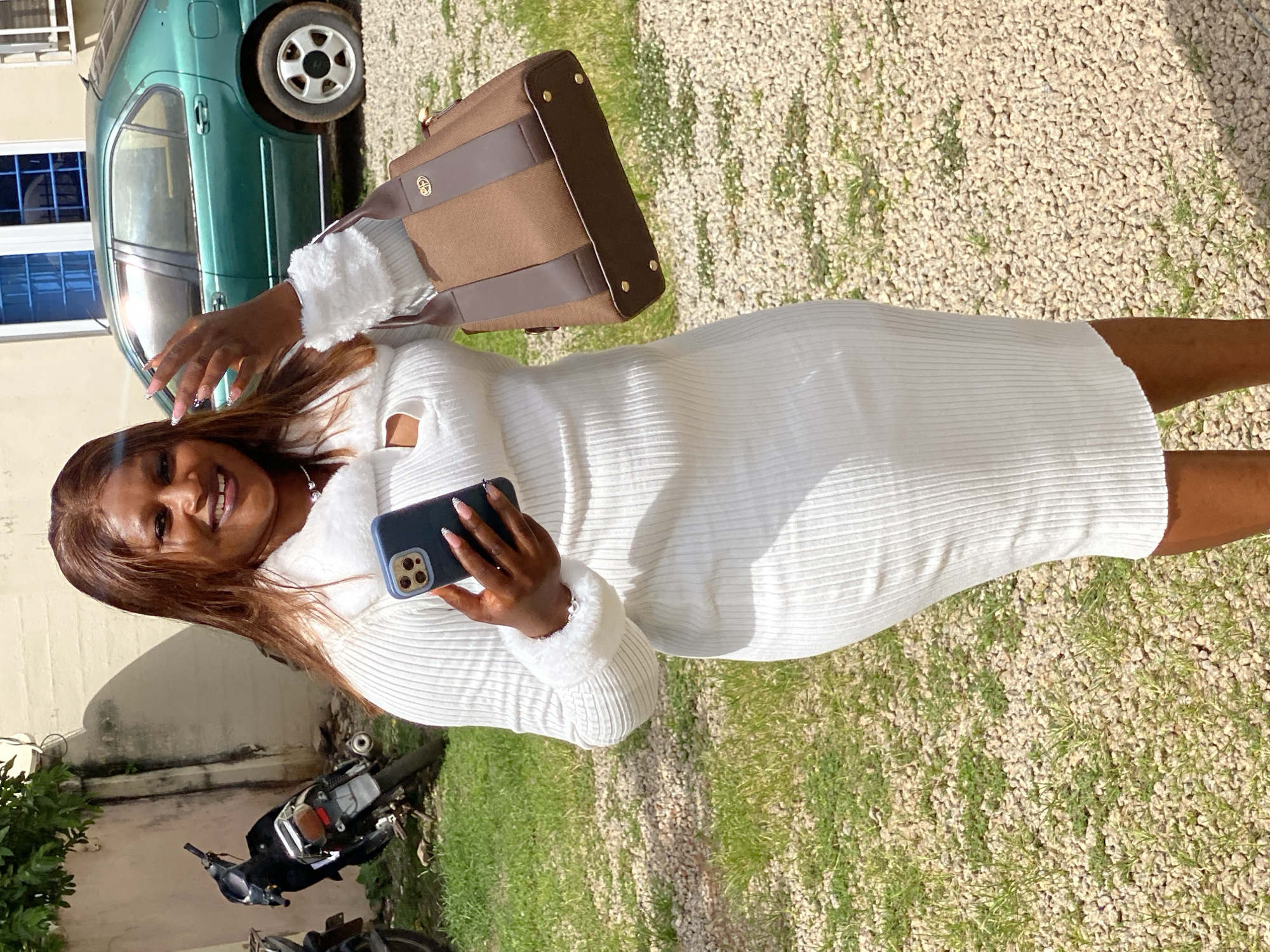 A woman, with long brown hair and a bright smile, is posing for a photo outdoors. She is wearing a white ribbed dress with fluffy trim on the sleeves. In her right hand, she holds a dark blue smartphone, which she uses to take a selfie. She is also carrying a brown and dark brown handbag over her left shoulder.
Behind her, a green car is parked on a gravelly surface. The car appears to be an older model. A motorcycle is visible in the lower left corner of the image, parked on grass. The background suggests a residential or outdoor area, with a light-colored building to the left and green vegetation. The lighting indicates it is daytime, likely sunny, with shadows cast on the ground. The overall mood appears cheerful and casual. No visible text is present in the image.