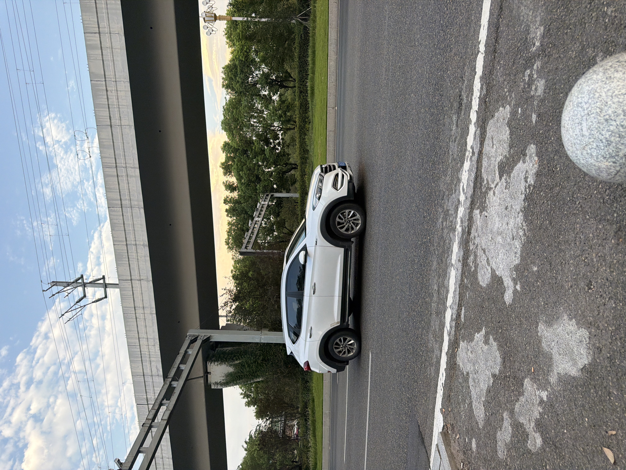 A white SUV drives on a paved road with a white lane marker. Overhead, a concrete overpass supports train tracks and power lines. Trees and greenery line the side of the road. In the foreground, a granite bollard is partially visible. The sky above is partly cloudy. The scene appears to be in Jiangjin, China.