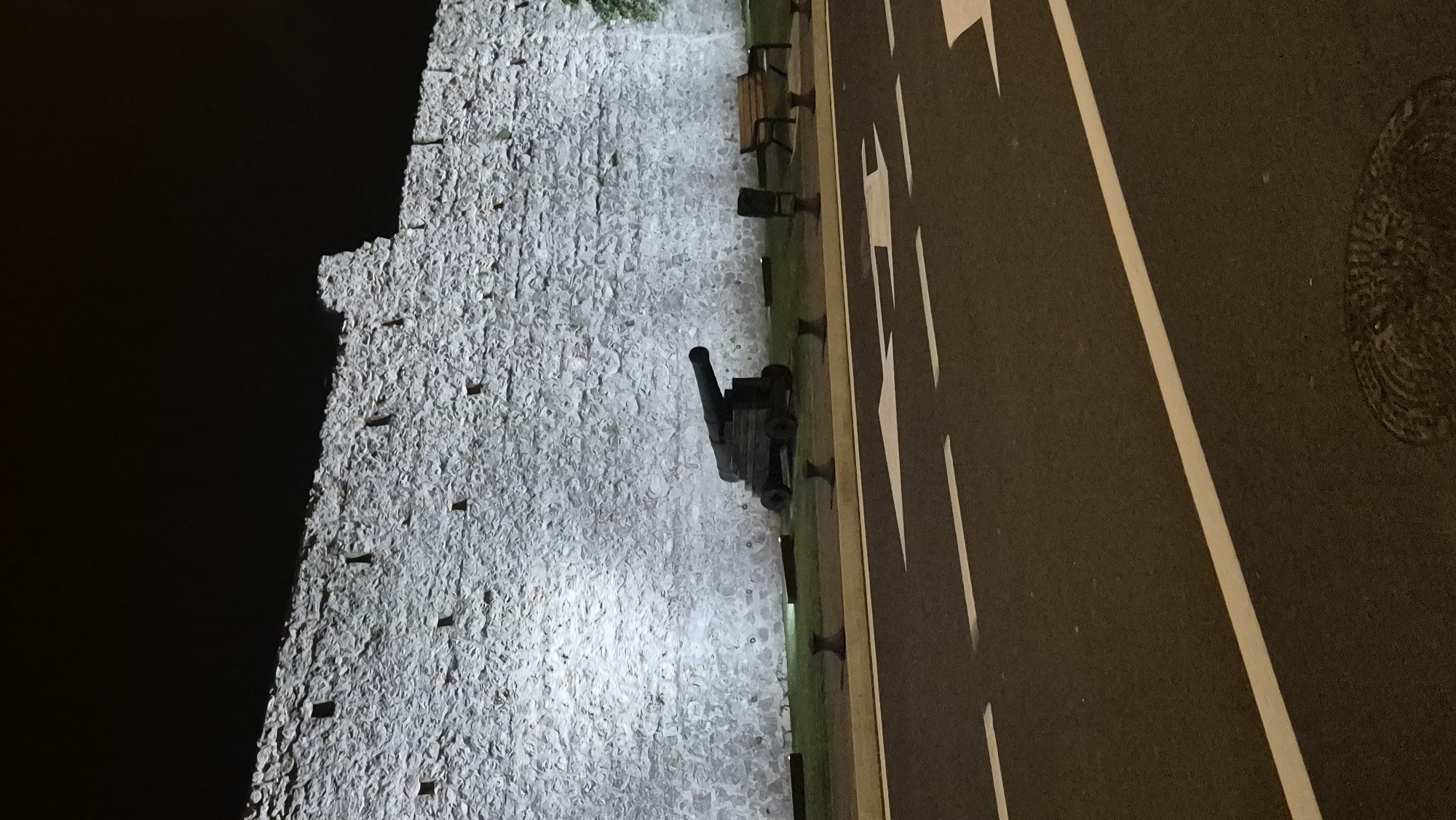 The image depicts a night scene of a stone wall illuminated by floodlights. In front of the wall, a historic cannon is displayed on a grassy area, accompanied by a park bench and a trash receptacle. A road with white lane markings and directional arrows is in the foreground. The environment is dark, with the exception of the illuminated wall and the road. There are no people visible in the frame.