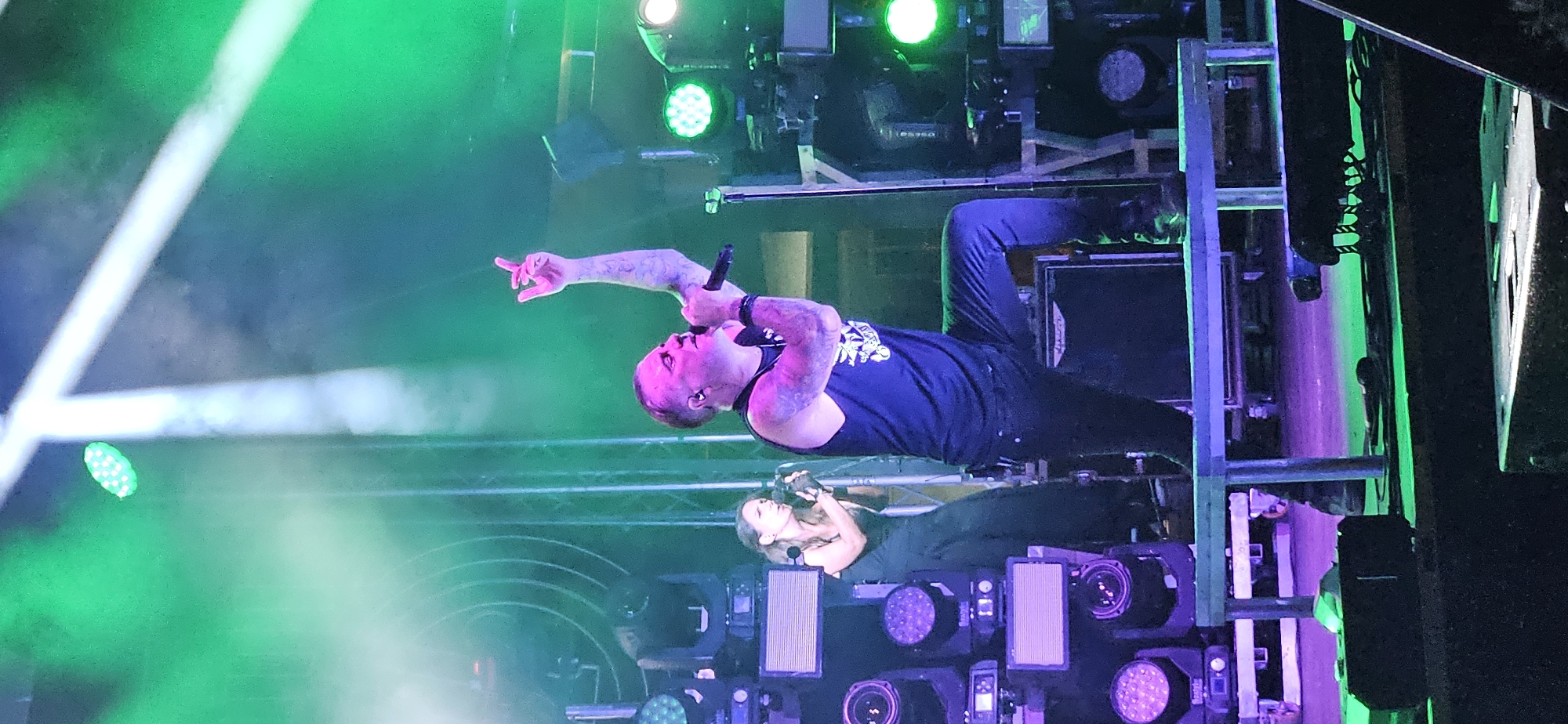 The image captures a live musical performance on a stage illuminated by vibrant green and white lights. In the foreground, a male singer, with visible tattoos on his arms, is center stage. He wears a black tank top and dark pants, holding a microphone and gesturing with his right hand. He is kneeling on a low platform. To his left and slightly behind him, a female singer in a dark dress holds a microphone and appears to be singing. The background is filled with stage lighting equipment, including multiple spotlights and speakers. The overall atmosphere suggests a concert or live music event. The image was taken in Hévíz, Hungary.