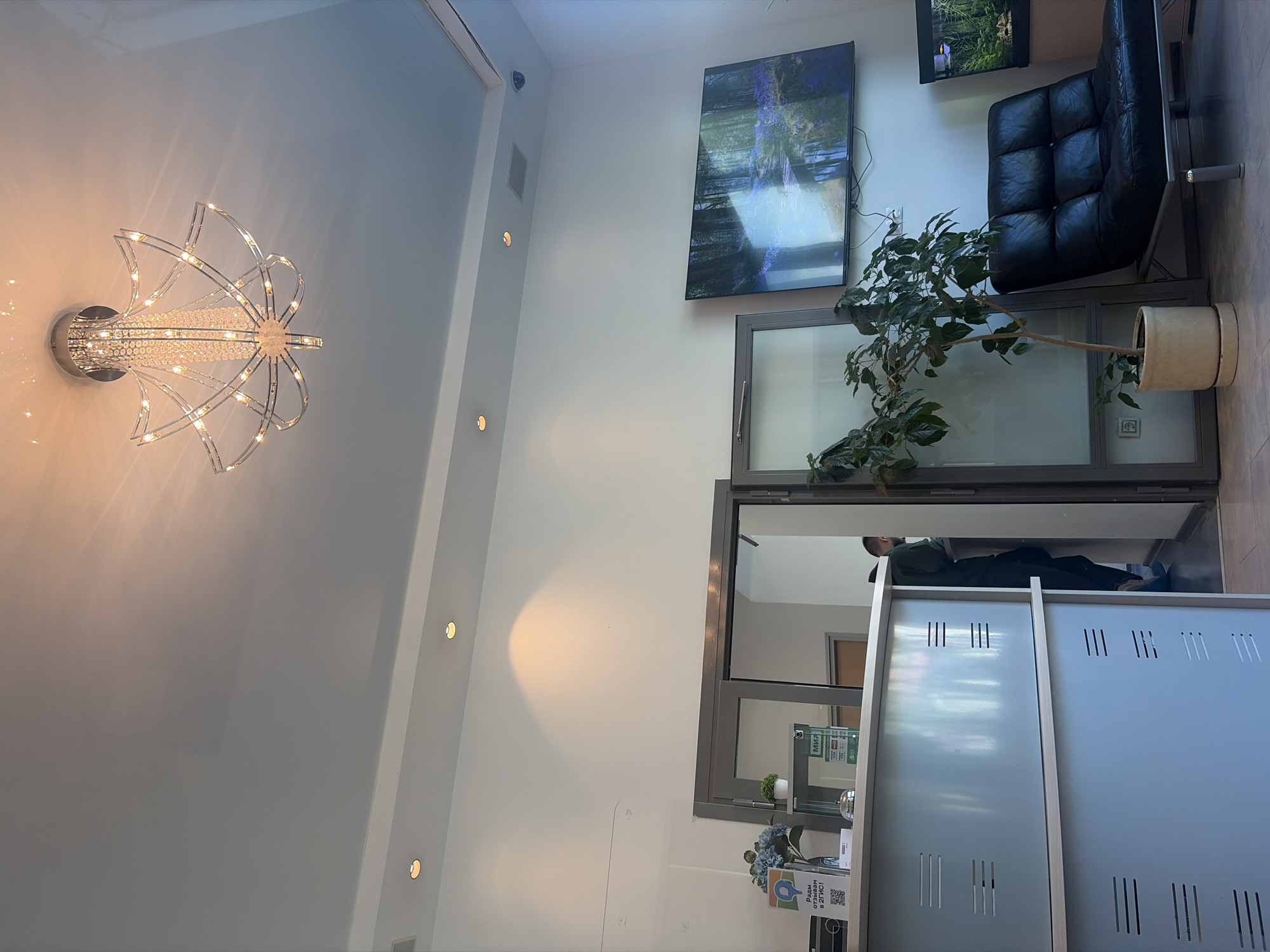 The image depicts the interior of a building in Saint Petersburg, Russia. A large, decorative chandelier hangs on the ceiling. On the wall are a large abstract painting, a television, and a black leather chair. A potted plant sits on a ledge near the window. A person is lying down and appears to be sleeping near a white cabinet or counter.