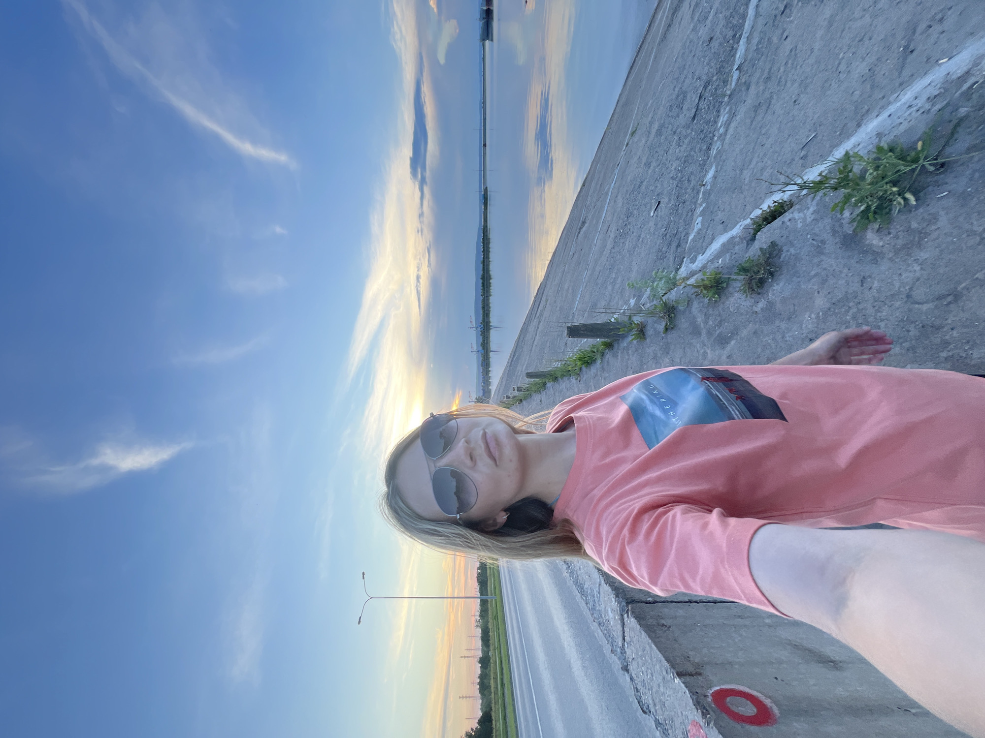 In Chaikovsky, Russia, a young woman with blonde hair and sunglasses takes a selfie on a concrete embankment. She is wearing a coral-colored t-shirt with a picture on it. To her left is a road with a streetlight. To her right is a body of water reflecting the blue sky and clouds. The sun is setting, casting a warm glow over the scene.