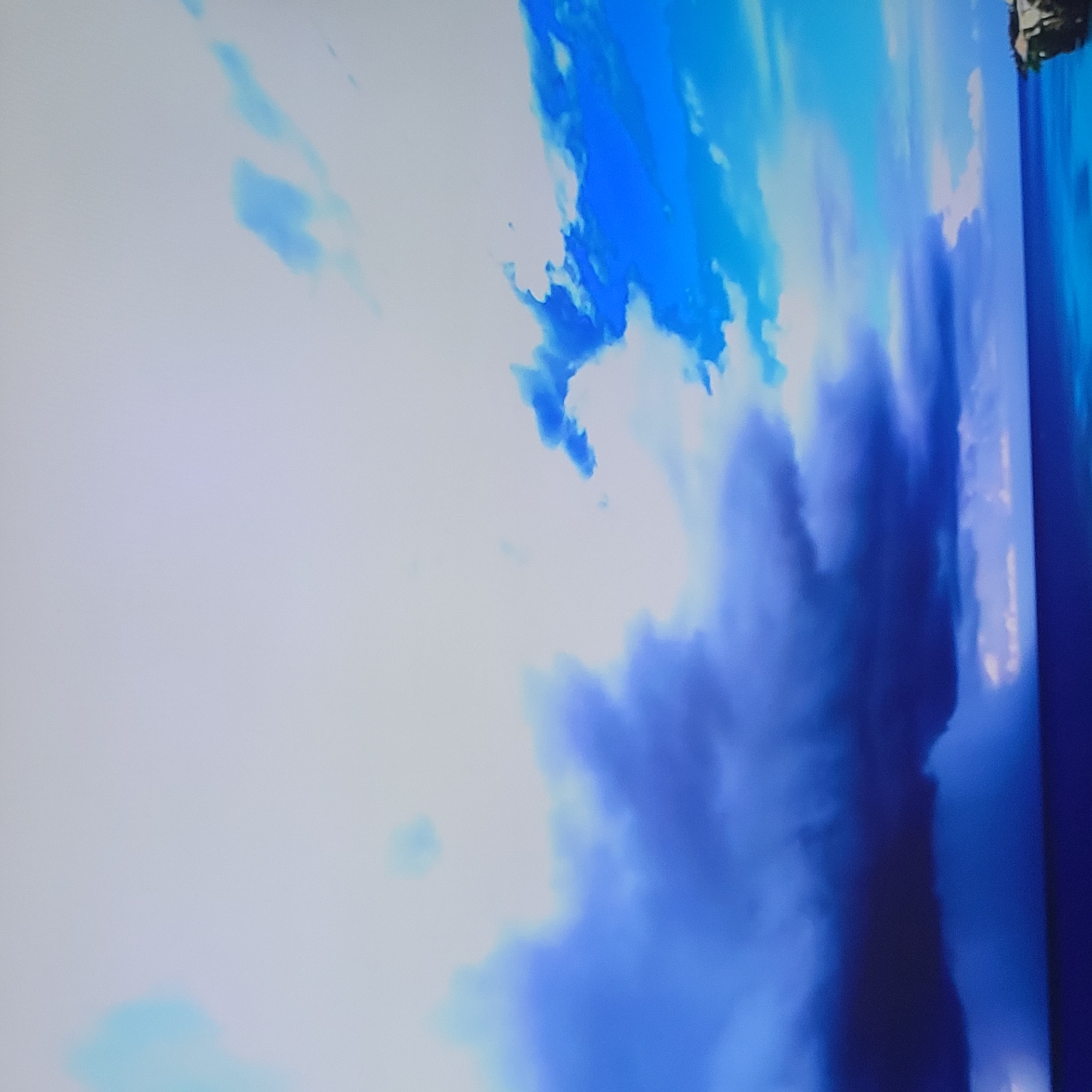 The image depicts a cloudy sky with varying shades of blue and white. Darker blue hues form cloud formations, while lighter blues and whites create a brighter background. In the top right corner, there is a blurred hint of greenery, possibly foliage. The scene appears to be a section of a larger image or display, potentially depicting a landscape with the sky as a prominent feature. Yerevan, Armenia, is not discernible in the image.