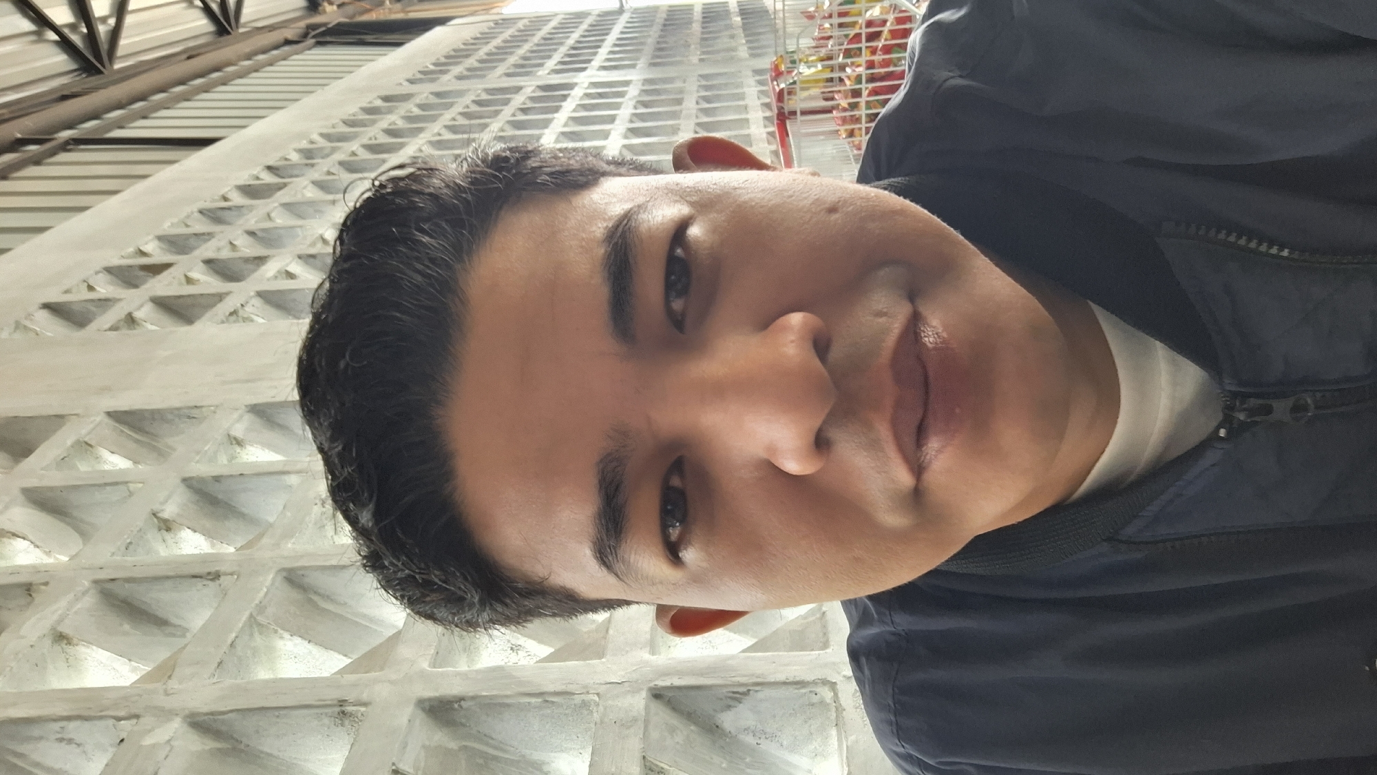 A person is standing near a building with a geometric facade. The person has dark hair, brown eyes, and is wearing a dark blue jacket over a white shirt. The building has a repeating pattern of triangular recesses. The location is plausibly Lhokseumawe, Indonesia, given the appearance and style of the building.