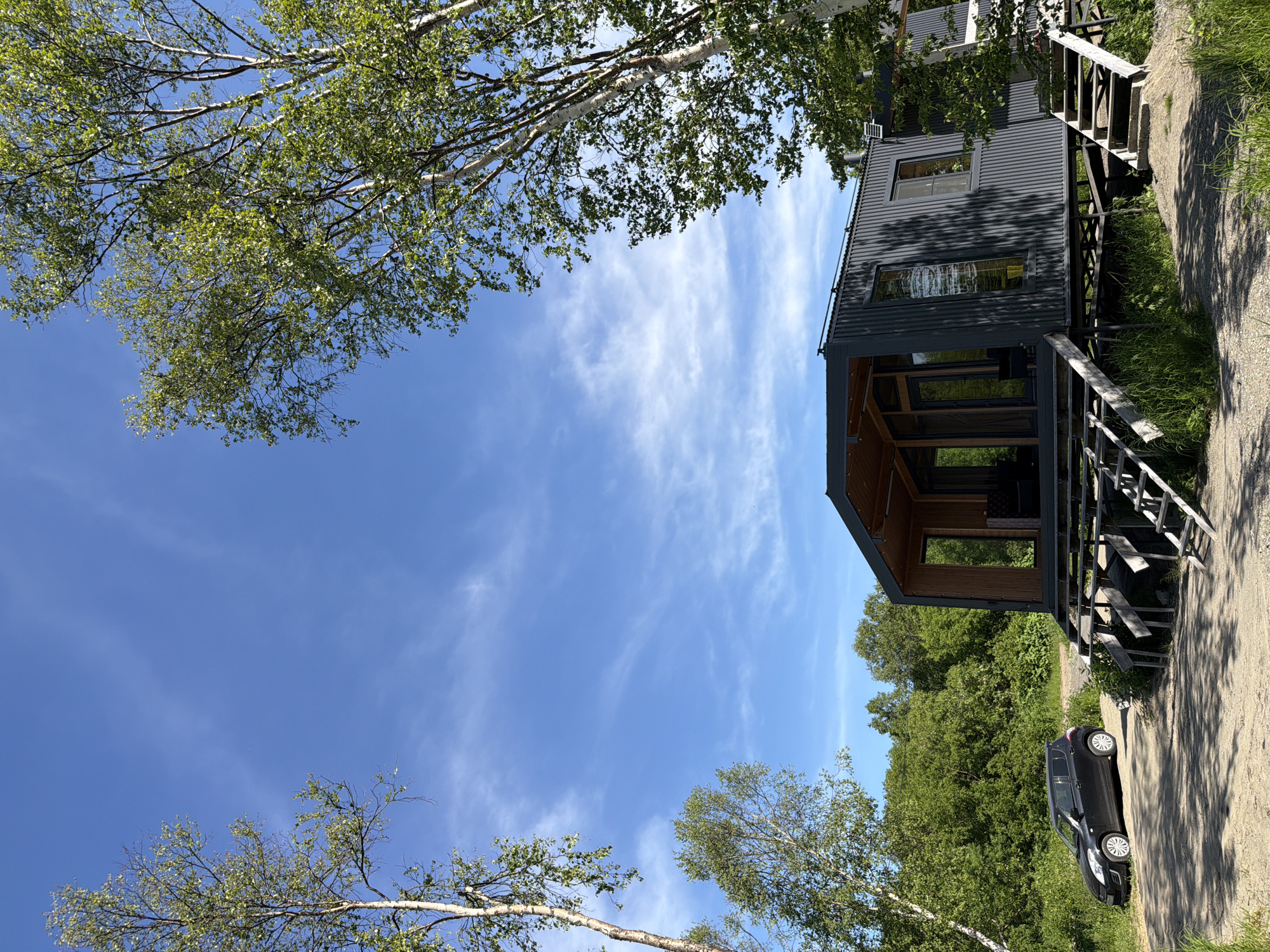 The image shows a contemporary, dark grey corrugated metal house situated on stilts in a natural, rural setting under a bright blue sky with scattered white clouds. The house has a modern design with large glass windows and an open-concept porch area. Two slender birch trees with light bark and green foliage flank the house, one on the left and one on the right, framing the structure. In the foreground, a black compact car is parked on a dirt path to the left of the house. The environment appears to be a wooded area with lush green trees and grass surrounding the property. The location is identified as the city of Novyy, Russia.