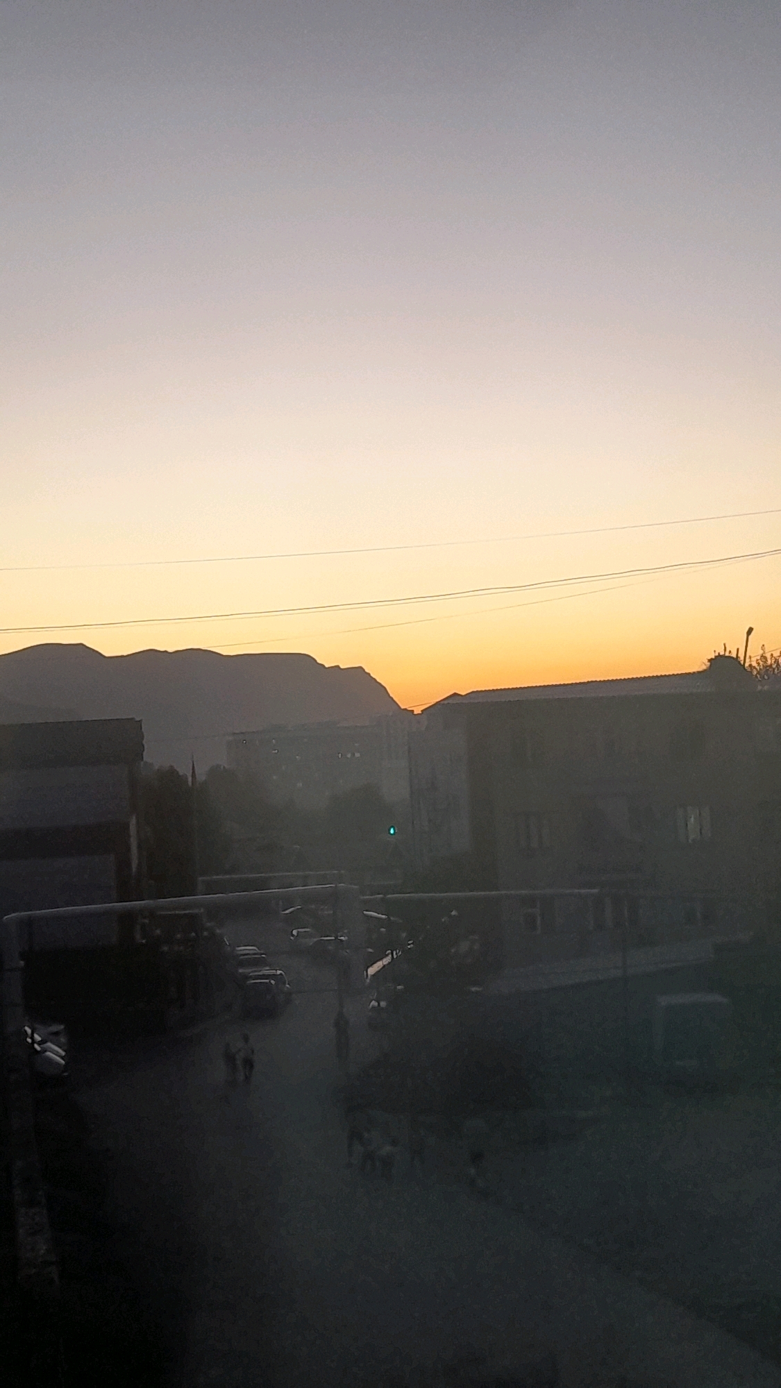 An exterior, eye-level, medium shot captures a dusky street scene in Osh City, Kyrgyzstan, with a sunset gradient of purple, orange, and yellow filling the sky. Silhouetted mountains form the backdrop. In the midground, several low-rise buildings, some appearing to be apartments or industrial structures, are visible. A road runs through the scene, with several parked vehicles, including what appear to be sedans and a van. A few individuals are depicted walking on the road. A traffic light, emitting a green signal, is also present. The overall lighting is dim, indicative of dusk or dawn.