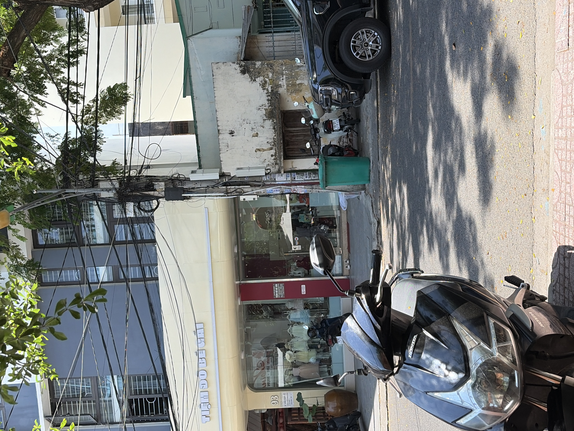 In the city of Nha Trang, Vietnam, a street scene unfolds under the dappled sunlight filtering through tree foliage. The foreground is dominated by a black scooter, its front end angled towards the viewer, revealing the Honda logo on its fairing. Behind the scooter, a dark-colored SUV is parked on the right side of the frame, its front wheel and fender prominently visible. The street is paved and appears to be in a commercial area, with a sidewalk of patterned bricks running along the bottom edge.
To the left of the parked vehicles, a shop with a light-colored facade and large glass windows displays children's clothing on mannequins. The shop's name, "TIỆM ĐỒ BÉ GÁI", is visible in white lettering above the entrance. A large brown ceramic pot with a plant sits outside the shop entrance. Further to the left, a part of a building with gray walls and numerous windows is visible.
Adjacent to the shop, an older, weathered building with a light-colored, peeling facade stands. A section of this building has a dark wooden door and a small, barred window. A green trash bin is placed near this older structure. The background features modern buildings with multiple stories and windows, and a complex network of electrical wires hangs overhead, crisscrossing the scene. The overall impression is one of a bustling urban environment with a mix of commercial activity and residential or office buildings.