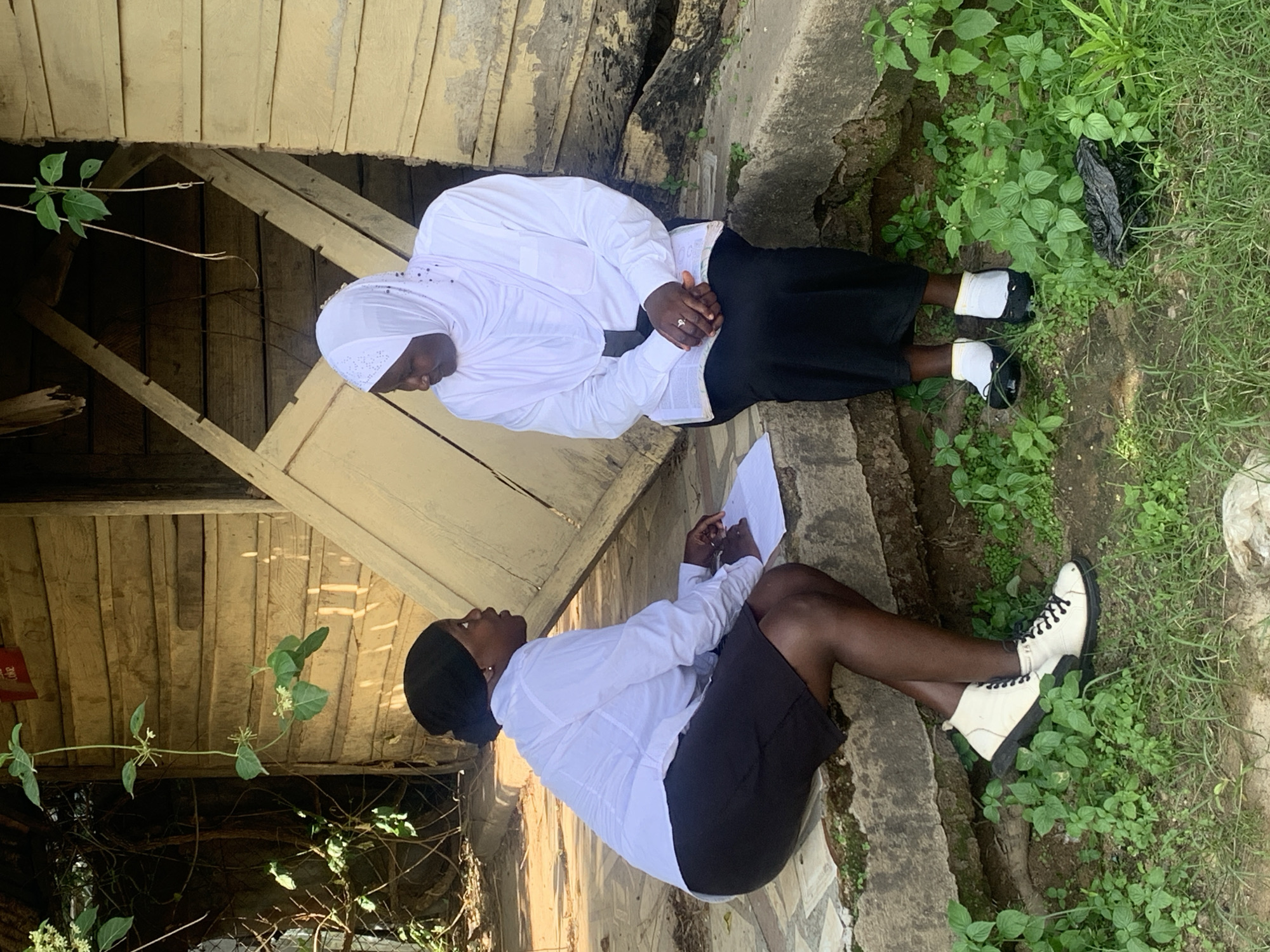 This image, captured in the city of Jos, Nigeria, shows two women interacting outdoors. A younger woman, of dark complexion, with a black head covering, a white long-sleeved shirt, and a black knee-length skirt, is seated on the left edge of a concrete step. She holds a white piece of paper in her lap and looks towards the second woman. Her footwear consists of white high-top sneakers with black laces. Seated to her right on the same concrete step is an older woman, also of dark complexion, wearing a white hijab, a white long-sleeved shirt, and a black long skirt. She holds an open book or document in her lap, with her hands clasped over it, and wears a ring on her right ring finger. Her feet are clad in black slip-on shoes with white socks. Both women are facing each other, indicating a conversation or interview.
The background features a weathered, faded yellow wooden building constructed with horizontal planks, showing signs of disrepair. A wooden door, partially ajar and appearing damaged, is visible behind the older woman. A red and white "Coca-Cola" sign is partially visible above the door frame. Overhanging vegetation, including a vine with green leaves and small white flowers, is present in the mid-ground. The ground level comprises dirt and patchy green foliage, with a black plastic bag resting near the older woman's feet. The scene is illuminated by natural daylight.