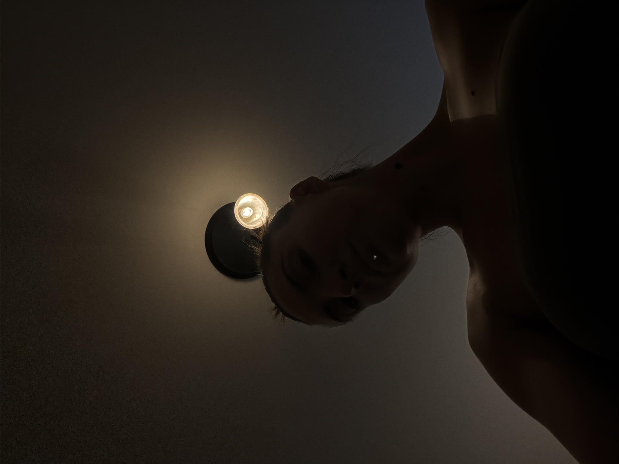 The image, taken from a low angle looking upwards, features a person illuminated by a ceiling light. The person's face is partially visible, with dark hair pulled back. They appear to have a piercing on their lip. Their bare shoulder and upper chest are also visible, with shadows obscuring much of their form. The background is a dark, plain ceiling. There is no indication of the city of Sharm El Sheikh, Egypt, or any other specific location. The image appears to be a self-portrait or a candid shot taken indoors.