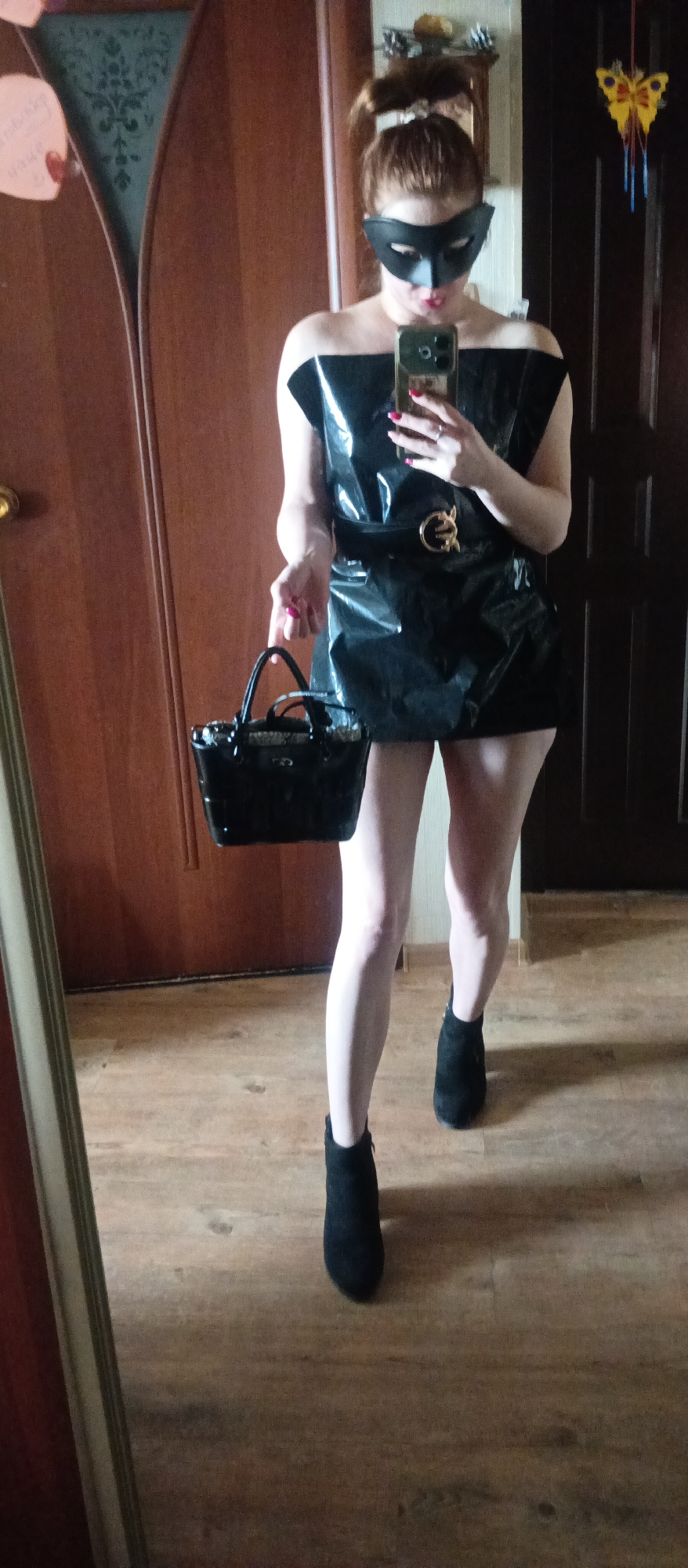 A woman wearing a black dress, a black mask, and black ankle boots is standing indoors. She is holding a black handbag in her left hand and a smartphone in her right hand, seemingly taking a photo in a mirror. The background shows wooden doors, a wooden floor, and some decorations on the wall. The location is Unknown, Unknown.