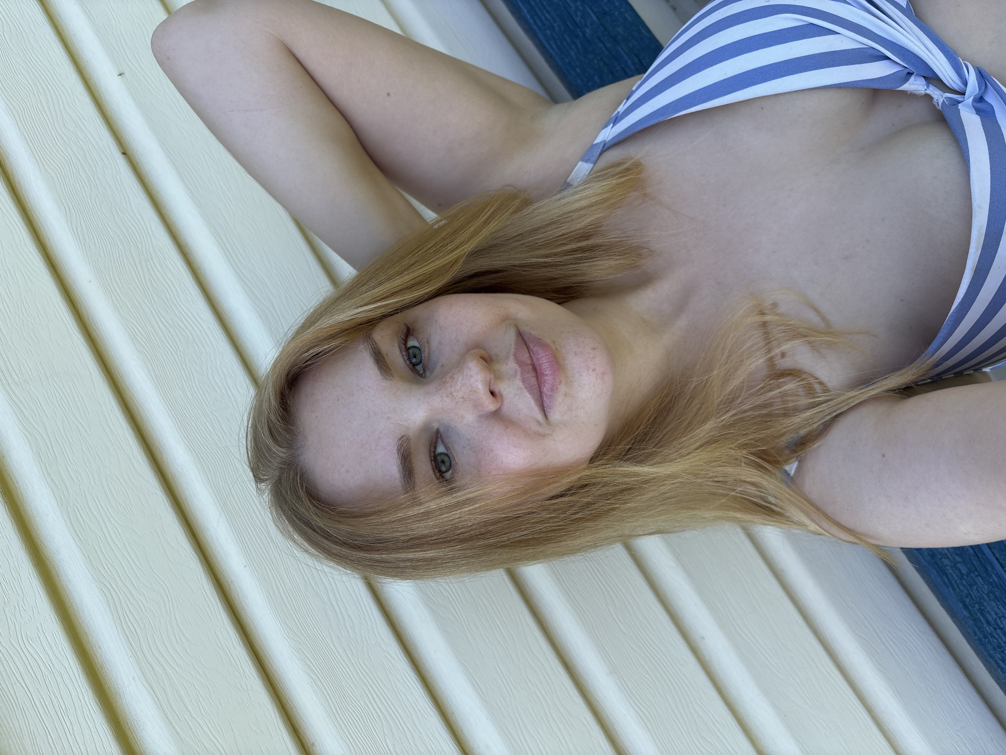 A young woman with blonde hair and fair skin is captured in a close-up selfie. She is wearing a blue and white striped bikini top. Her left arm is raised, resting behind her head, and she is looking at the camera with a slight smile. The background consists of light-colored horizontal siding, with a dark blue stripe visible at the bottom. The scene appears to be outdoors, possibly on a bench or seating area, with the siding suggesting a building structure. The image is taken from a high angle, looking down at the woman. No specific location in the city of Дунилово, Russia, can be identified from the visual information provided.