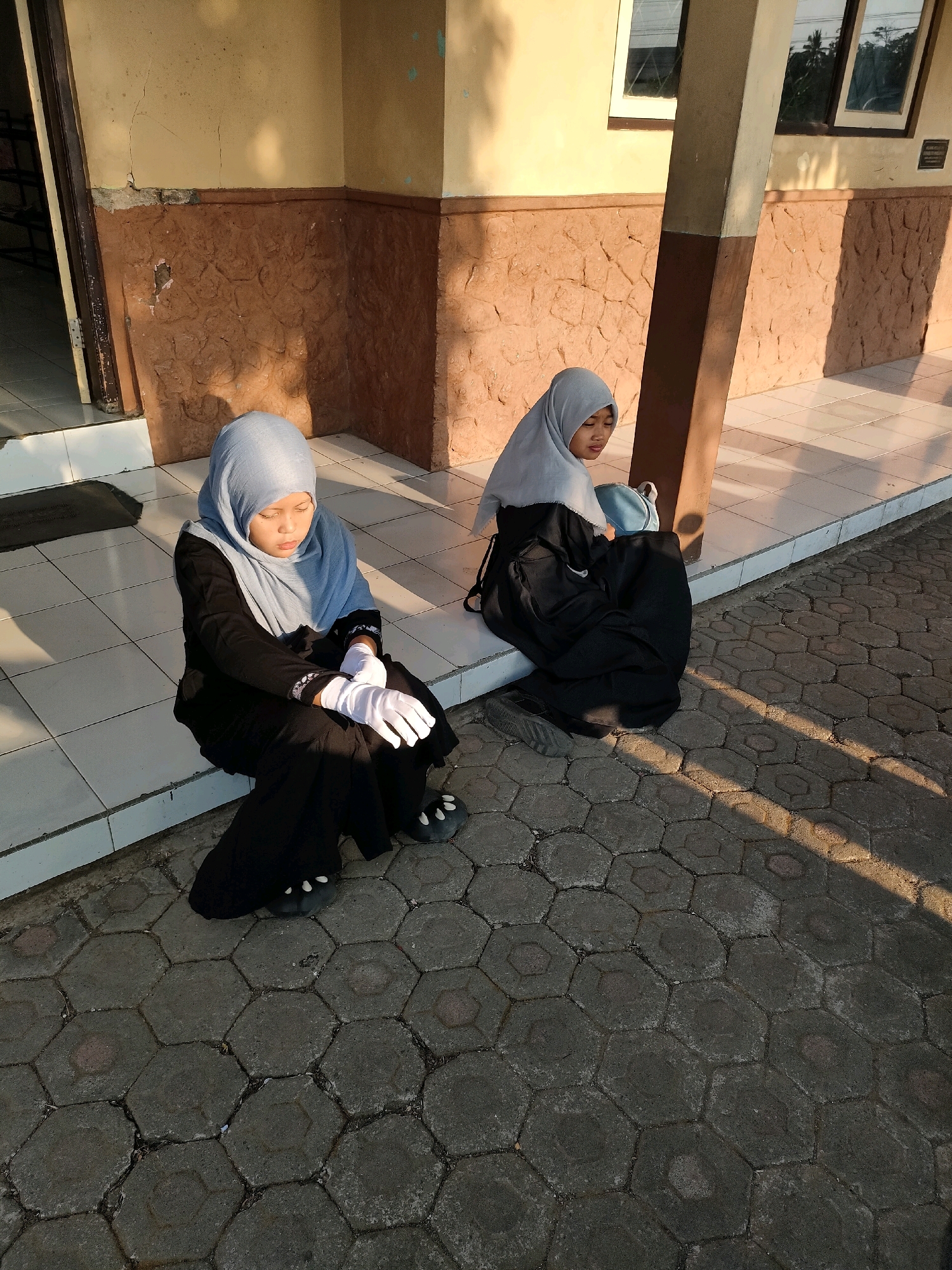 Two young girls wearing hijabs are sitting on the ground next to a building. The girl in the foreground is wearing a light blue hijab and a black dress, with white gloves on her hands. The girl in the background is wearing a light blue hijab and a dark dress, and has a light blue backpack next to her. The building has a textured brown lower wall and a yellow upper wall. The ground is paved with hexagonal stones. The scene appears to be outdoors during the daytime, with sunlight casting shadows. The location is identified as Bojong, Indonesia.