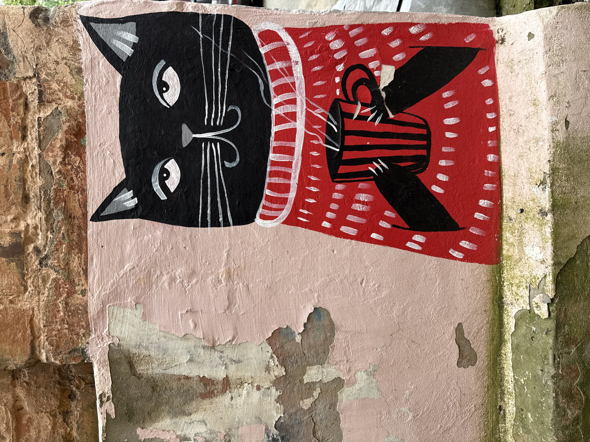 The image is a close-up photograph of a mural painted on a weathered, pink-colored concrete wall. The mural depicts a black cat wearing a red, high-necked sweater with white dashes. The cat has white eyes, white whiskers, and a grey nose. It is holding a red and black striped mug with steam rising from it. The concrete wall is cracked and peeling in places, revealing older layers of paint and a rough, textured surface underneath. Above the pink wall is a section of rough, reddish-brown brickwork. The bottom of the image shows more peeling paint and some green mossy growth on the lower concrete structure. The style of the mural is simplistic and cartoonish. The specific location in the city of Votkinsk, Russia is not determinable from the image itself.