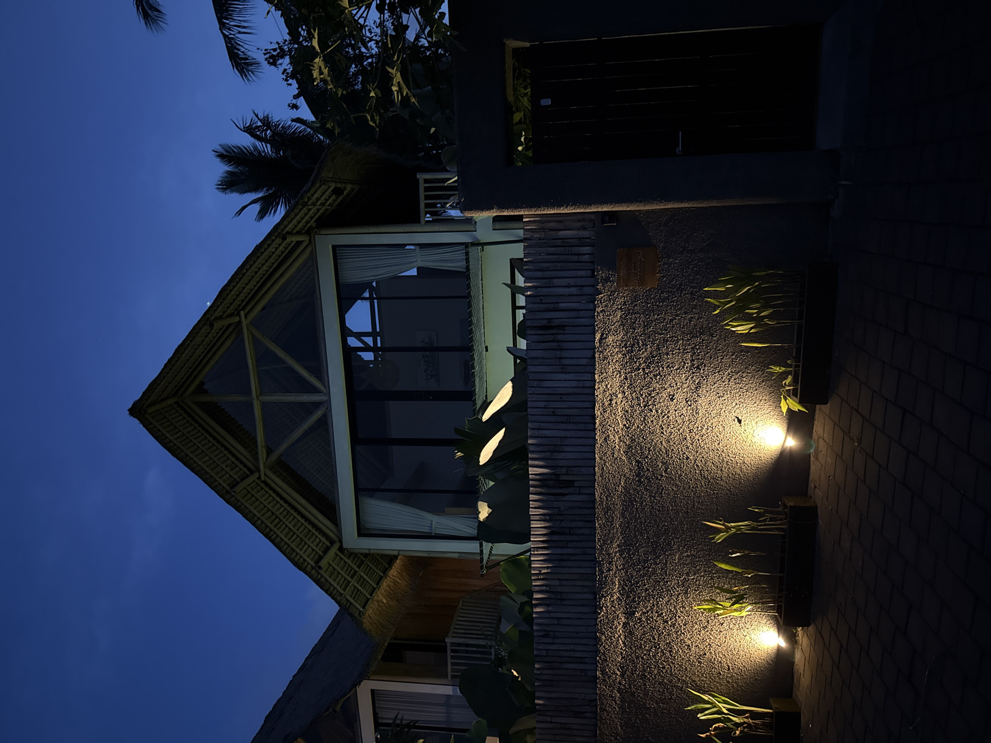 The image shows a building in Pejeng, Indonesia, at night. The building has a thatched roof and a triangular front facade with large windows. A bamboo wall is adjacent to the building, and plants are growing in containers along the wall, illuminated by lights. Palm trees are visible in the background against the dark blue sky. The ground is paved with brick.