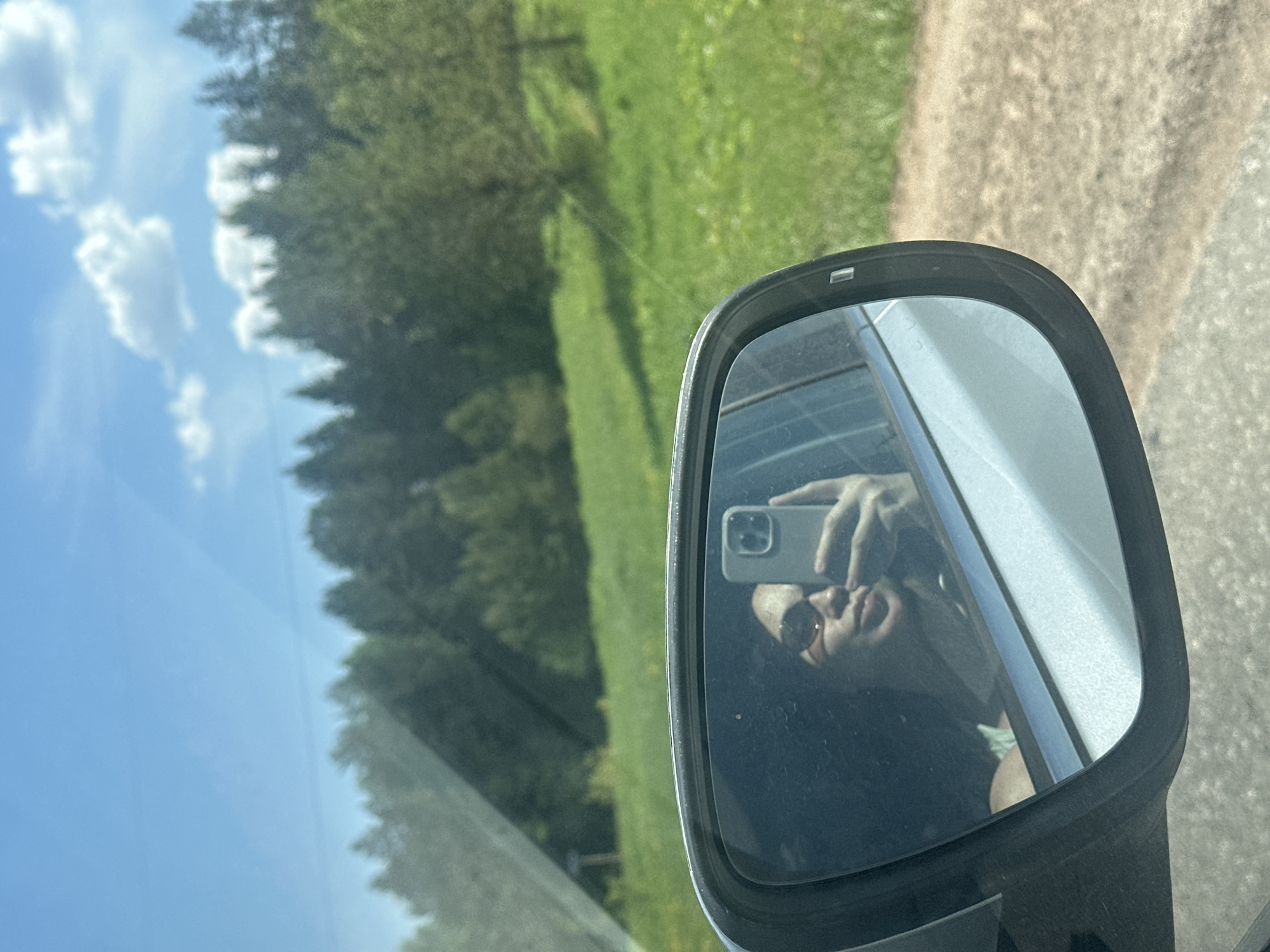 The image shows a car's side mirror, reflecting a person taking a photo with a smartphone. The person is wearing sunglasses. The mirror is attached to a car moving along a road. The background, visible through the car's window and partially reflected in the mirror, consists of trees, a green field, and a blue sky with clouds. The location is identified as being somewhere in the city of Staryi Chul'tem, Russia, based on the image's context (although this cannot be determined from the image itself).