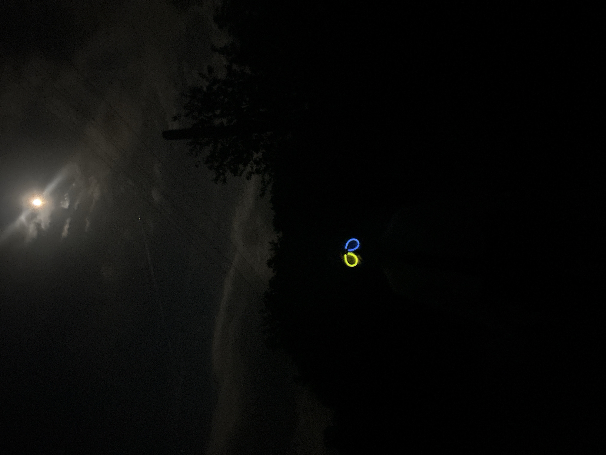 The image depicts a night scene in Konska, Bulgaria. In the left portion of the frame, a bright moon shines through clouds. To the right, power lines are visible adjacent to silhouetted trees. In the central-right portion of the frame, two glowing bracelets, one blue and one yellow, are discernible. The majority of the image is enveloped in darkness, obscuring details of the surrounding environment.