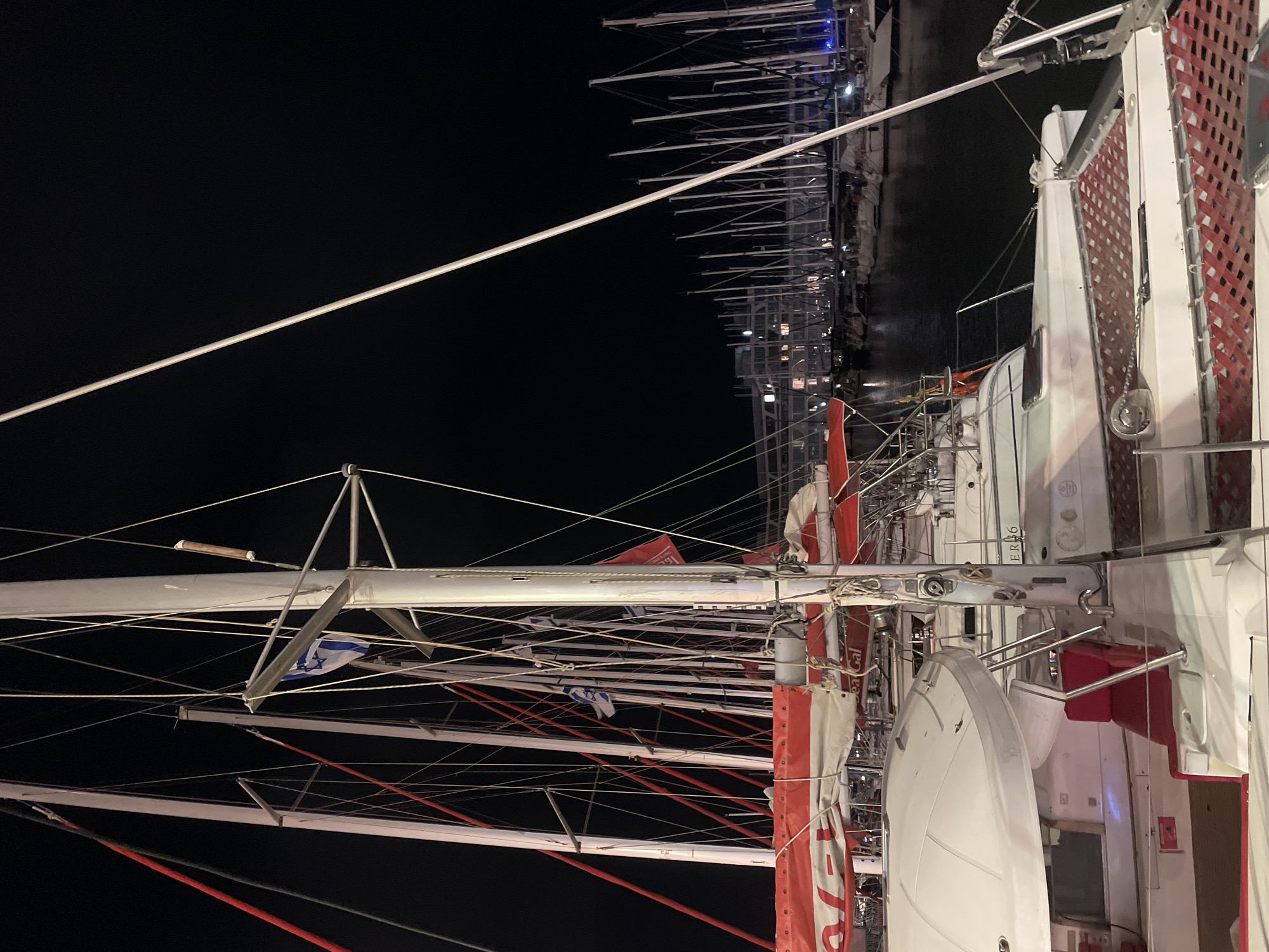 The image captures a night scene at the marina in Herzliya, Israel. A cluster of sailboats are docked, their masts creating a complex network of lines against the dark sky. Some masts are adorned with Israeli flags. The lighting suggests it is nighttime. The foreground shows the side of a boat with distinct features such as red and white detailing, a satellite dish, and signage. The marina area in the background is visible, with other boats and lights reflecting on the water.