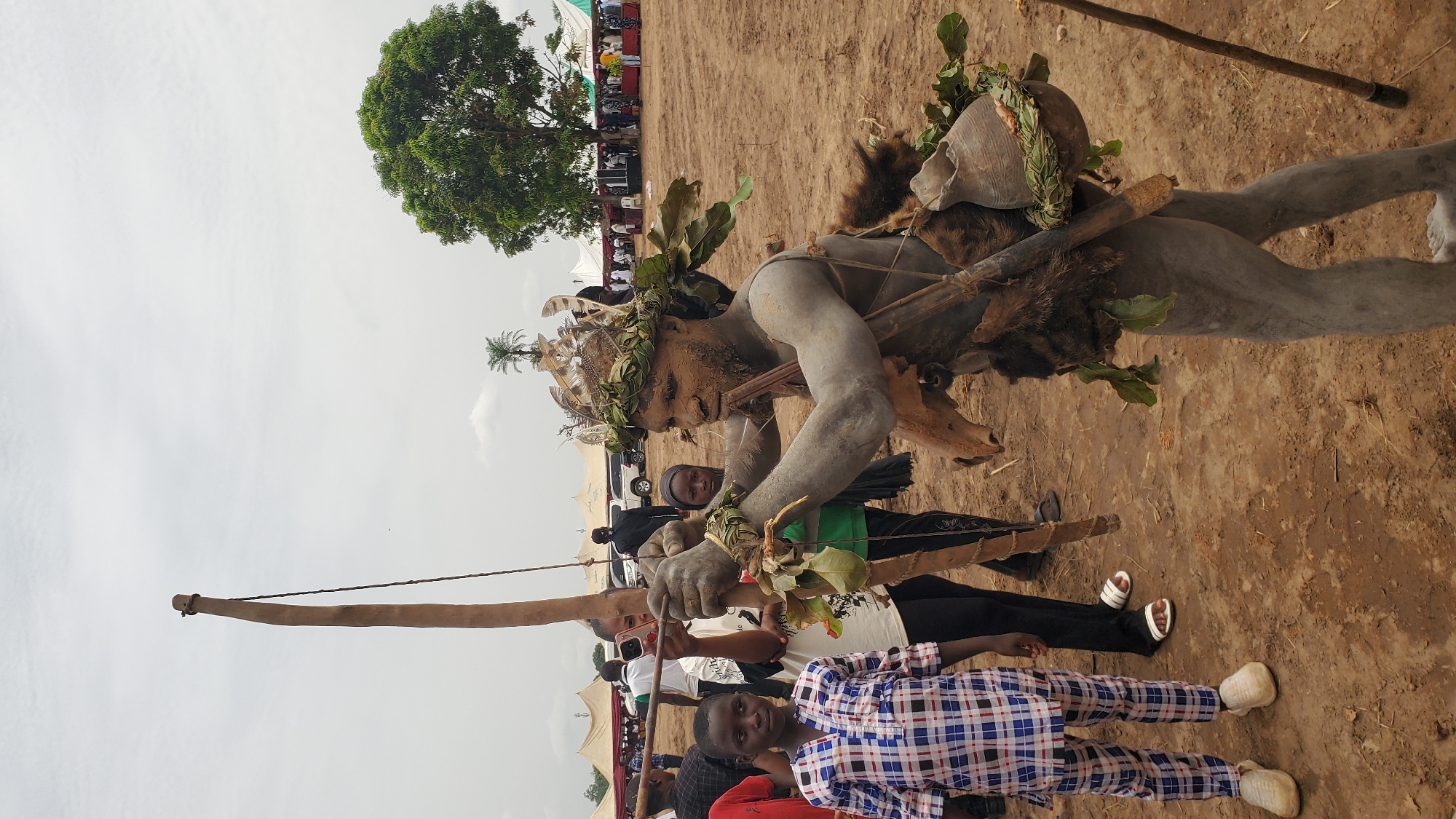 Akwanga, Nigeria: An elaborately decorated, life-sized mannequin (likely a representation of a spirit or deity) stands upright on a dirt ground. The mannequin is adorned with mud or clay, fur, leaves, and an animal skull. It holds a wooden bow. Several people, including a child in a blue and white plaid shirt, stand near the mannequin. Other individuals are partially visible in the background. Tents and vehicles are also present in the background, along with a tree and an overcast sky.