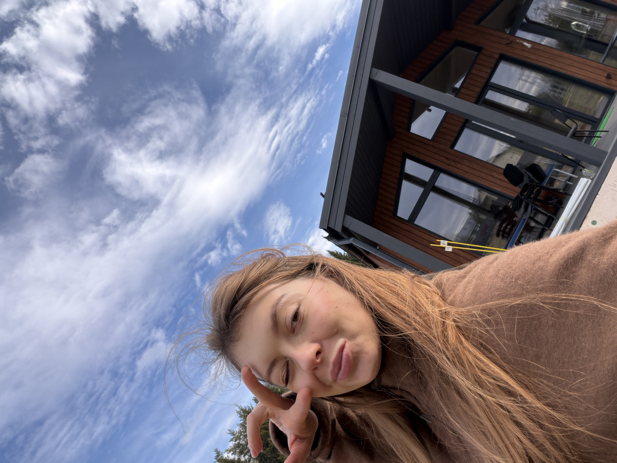 A young woman with light brown hair and wearing a brown sweatshirt poses for a selfie. She is winking with one eye and making a peace sign with her fingers near her face. In the background, there's a modern-style house with wood siding and multiple windows reflecting the sky. A blue sky with wispy clouds is also visible. The location is possibly in or around Zarech'ye-5, Russia.