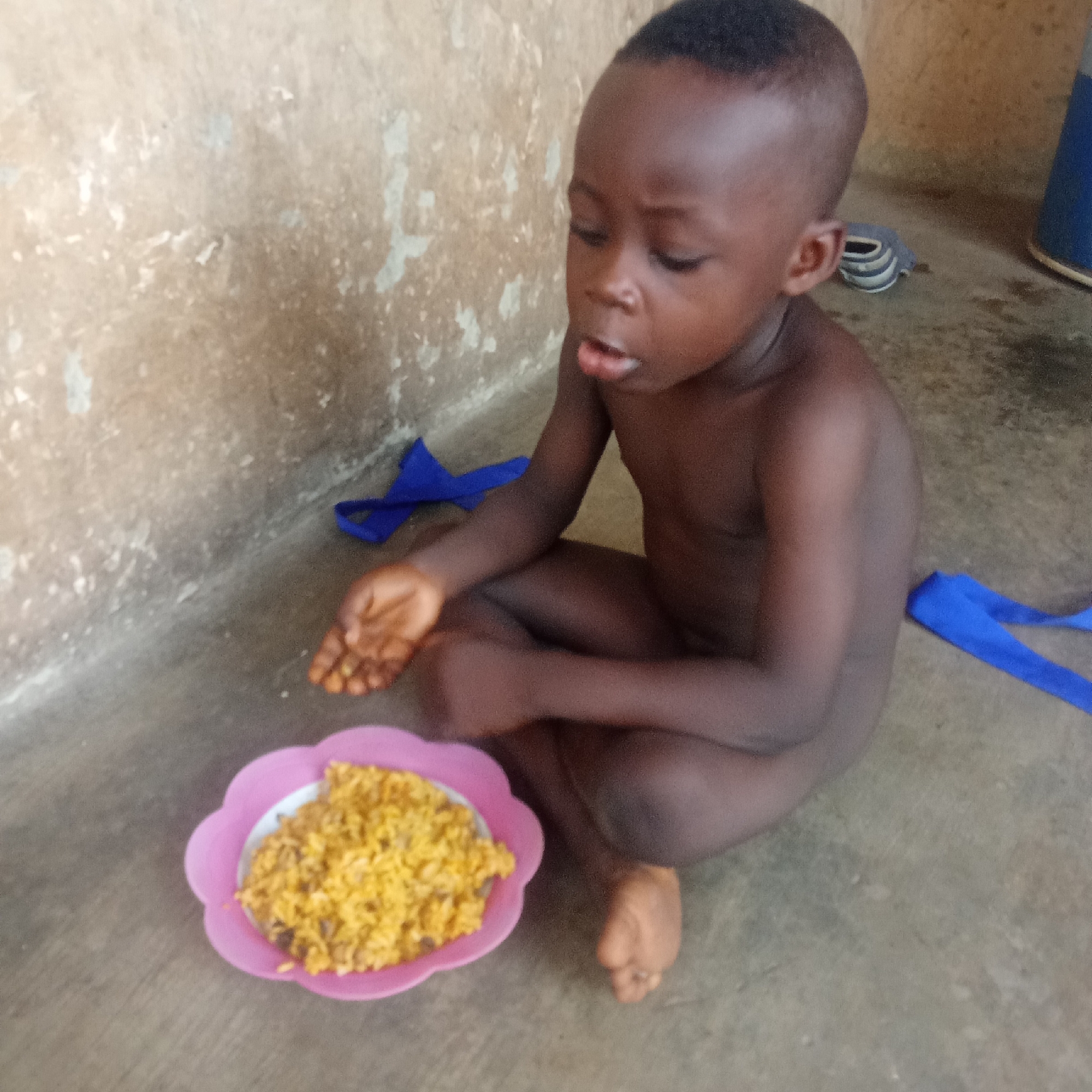 A young, dark-skinned child is sitting cross-legged on a concrete floor against a worn wall in what appears to be a residential setting in Akwanga, Nigeria. The child is shirtless and appears to be eating from a pink, flower-shaped plate filled with cooked rice or a similar dish. A blue ribbon or piece of fabric is nearby on the floor. A blue object, possibly a container, is visible in the background on the right. A pair of slippers are visible behind the child.