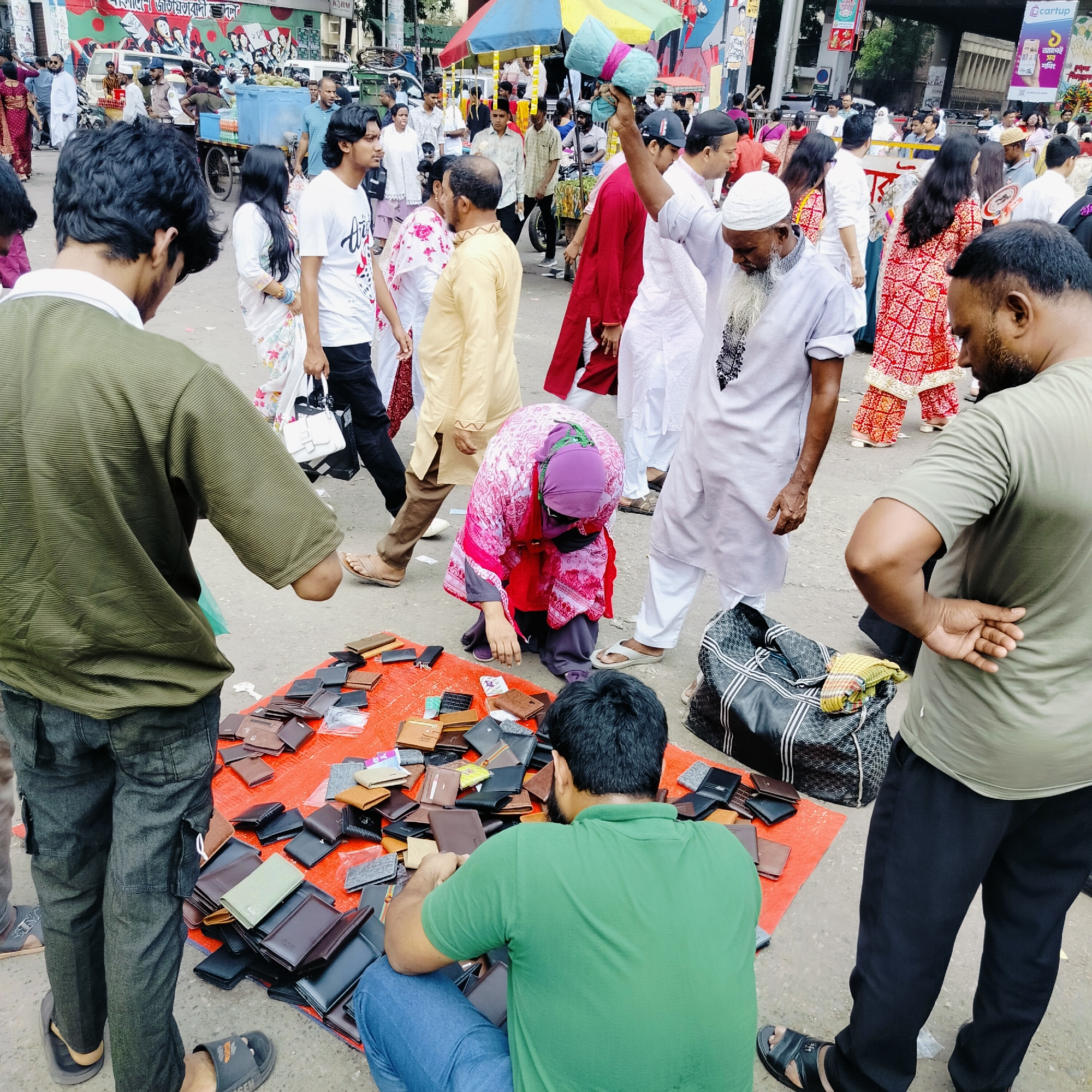 The scene is a busy street market in Dhaka, Bangladesh. In the foreground, a vendor sits on the ground surrounded by a large display of wallets on an orange sheet. Two men are examining the wallets, while a woman in a pink outfit kneels to look at them. A man in a white robe stands nearby, holding what appears to be rolled fabric. The background is filled with people walking on the street. A variety of shops and vehicles are visible in the distance.