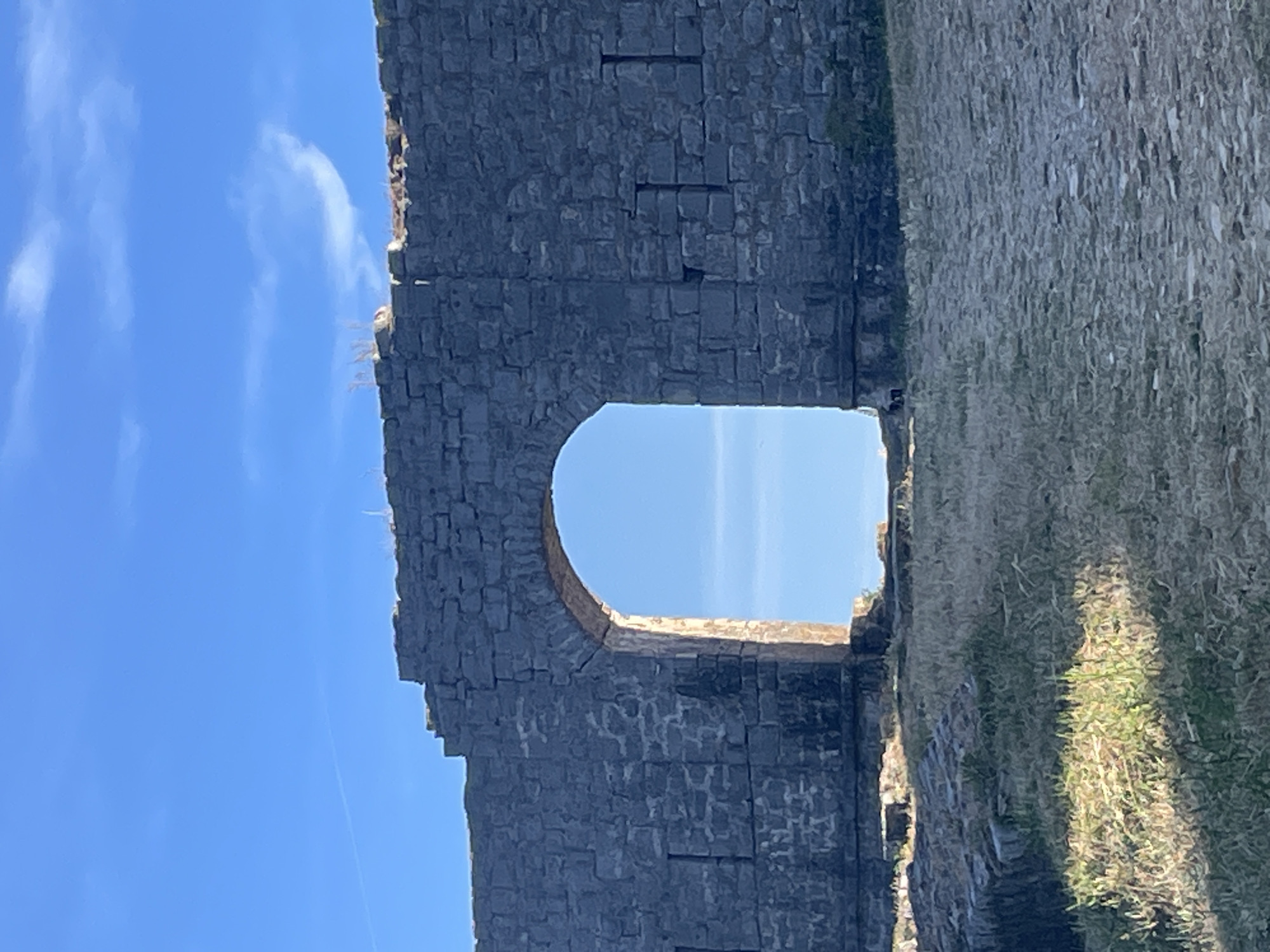 The image depicts a partial view of the Mogren Fortress, located in Budva, Montenegro. A stone archway is framed by the stone wall of the fortress, with a glimpse of blue sky visible through the opening. One side of the wall is covered in vegetation. The sky is mostly clear, with a few wispy clouds.