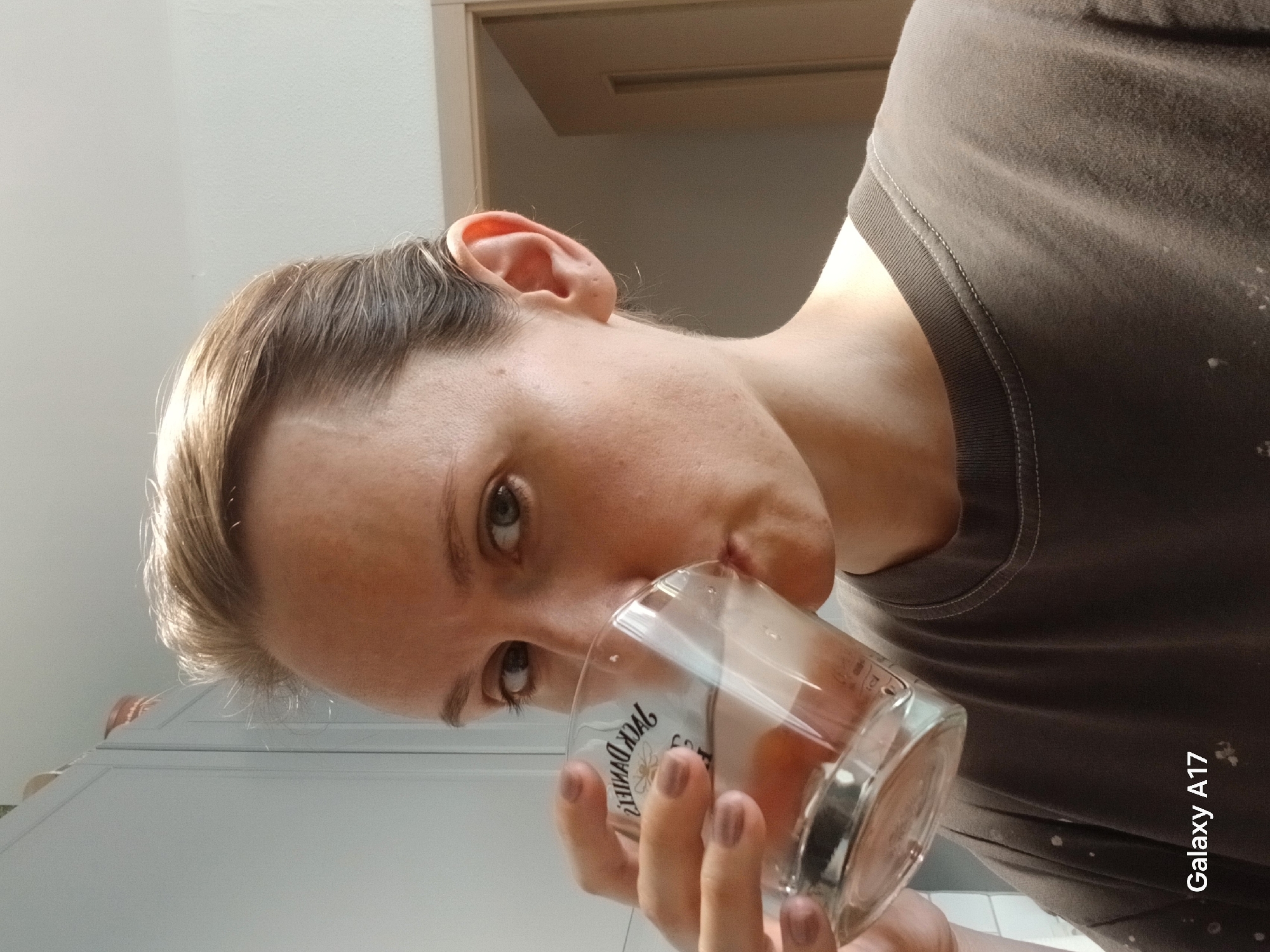 A person is drinking from a glass. The person is facing the camera and tilted their head. The glass is in their hand, and liquid is inside. The background includes a door frame, a counter, and a grey t-shirt. The location is unknown, and there is no indication it is Yekaterinburg, Russia. A "Galaxy A17" watermark is in the corner of the image.